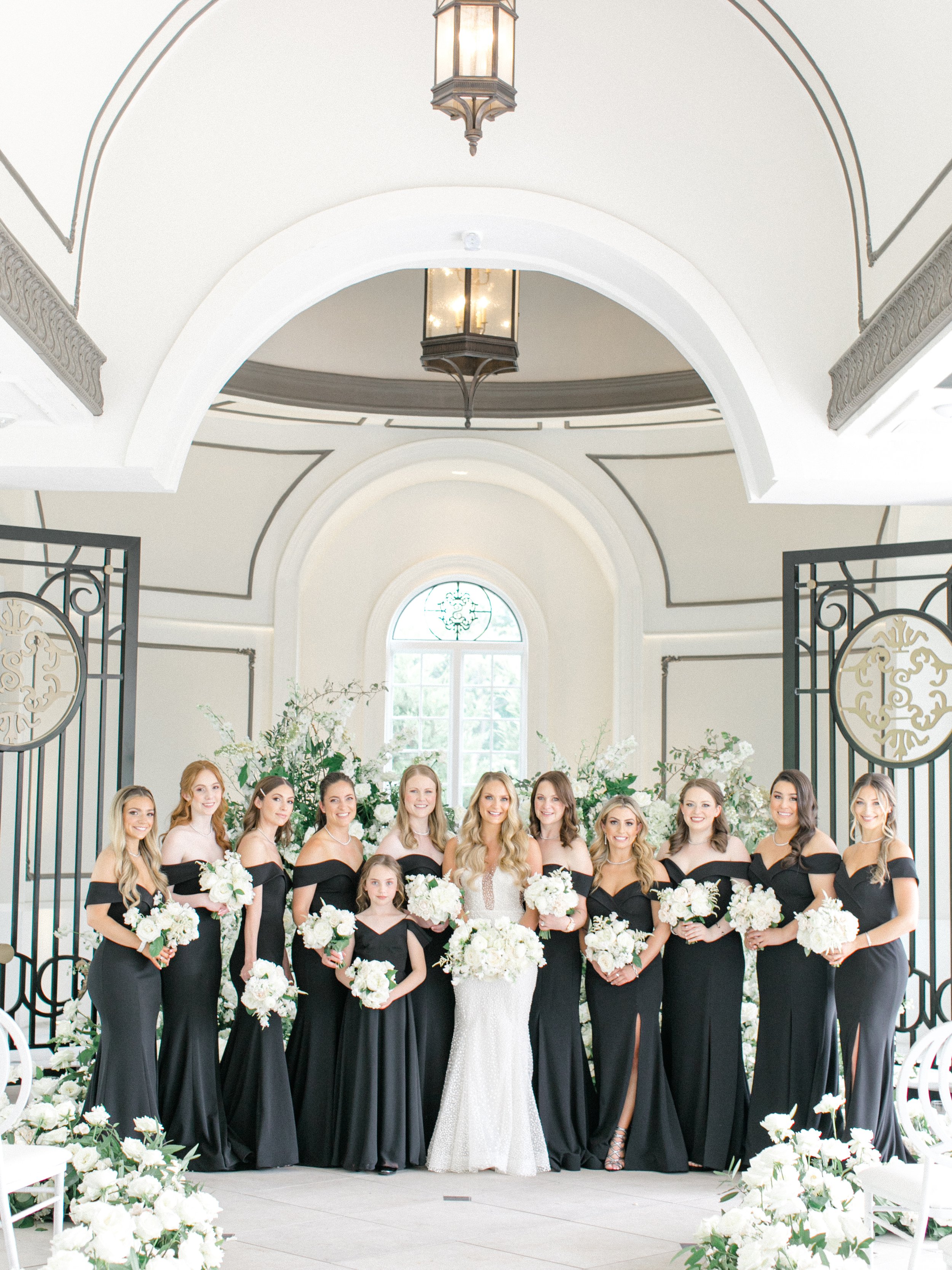 A bride and her bridesmaids standing inside a white hall with large windows, white floral arrangements, and decorative lighting. They are all dressed in black gowns and holding white bouquets.