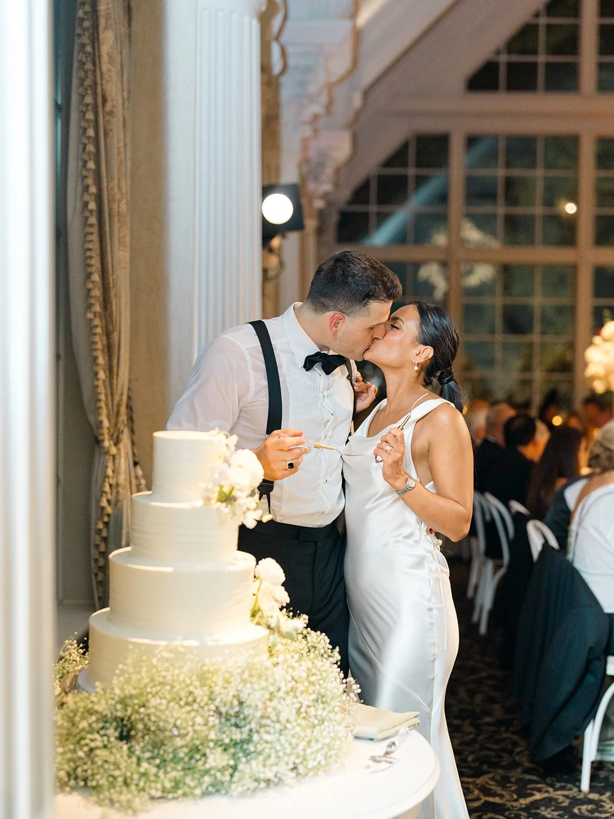 A newlywed couple shares a kiss during their wedding reception, standing near a multi-tiered wedding cake decorated with white flowers, in an elegant ballroom with large windows and guests seated at tables in the background.