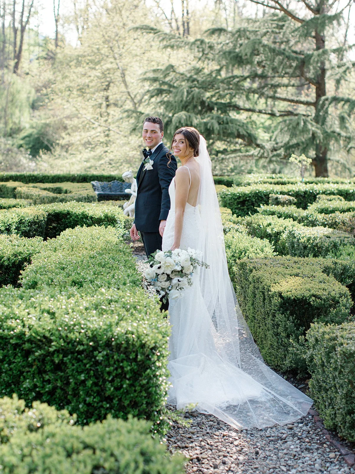 A bride and groom smile at each other in a garden, with the bride holding a bouquet of white flowers, surrounded by greenery and trees.