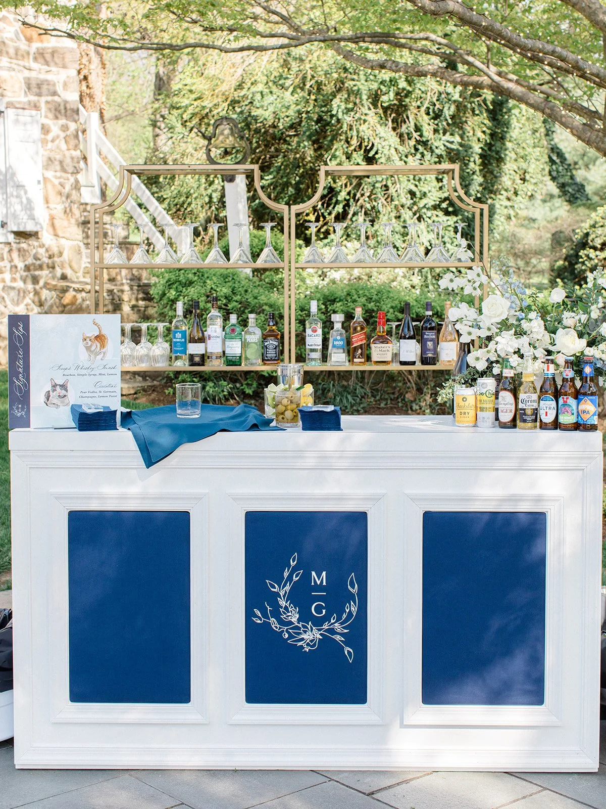 Outdoor bar setup with a white and blue bar counter, monogrammed with the initials M G and floral design, topped with various liquor bottles, beer bottles, garnishes, and a napkin, with a gold wine glass holder and glassware. Green trees and shrubs a
