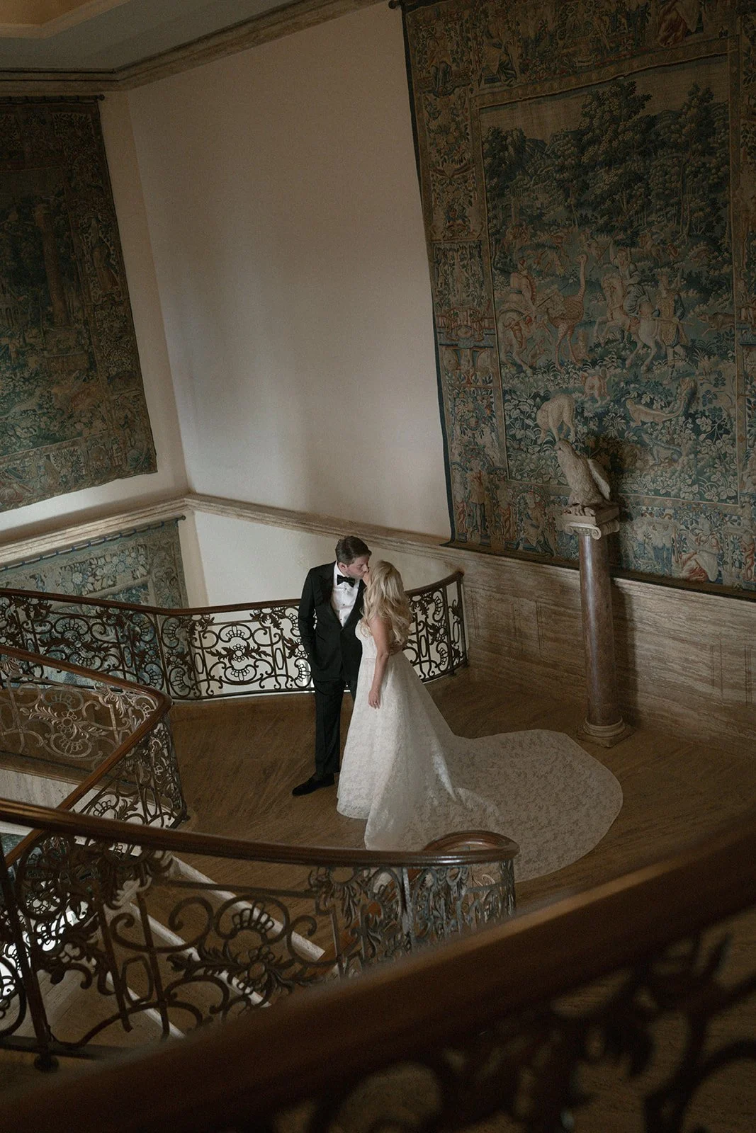 A bride and groom standing on a curved staircase, gazing at each other, in an elegant indoor setting with ornate tapestries and a column with a sculpture bust.