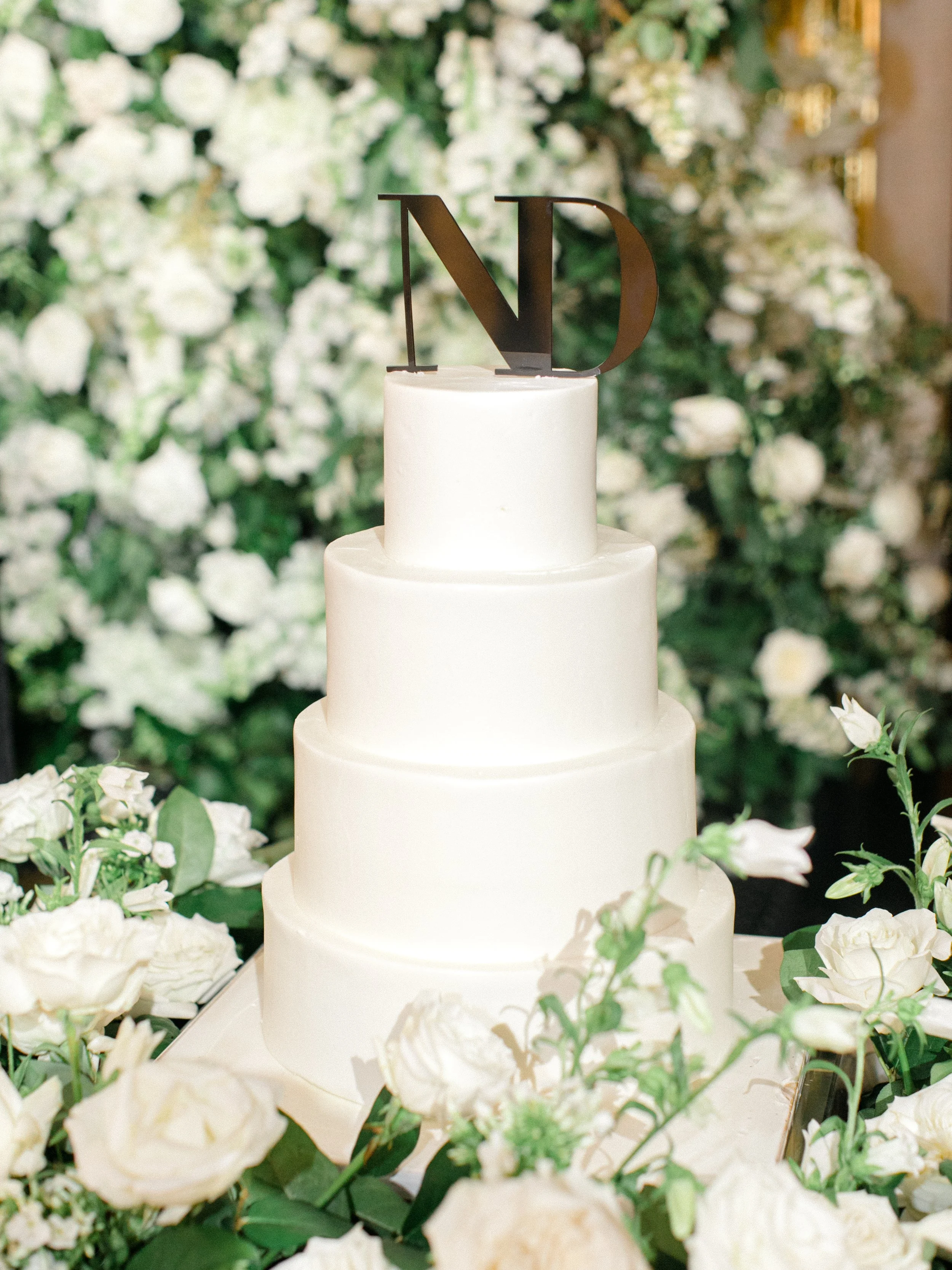 A four-tier white wedding cake with a black 'ND' cake topper, surrounded by white roses and greenery, against a backdrop of white flowers.