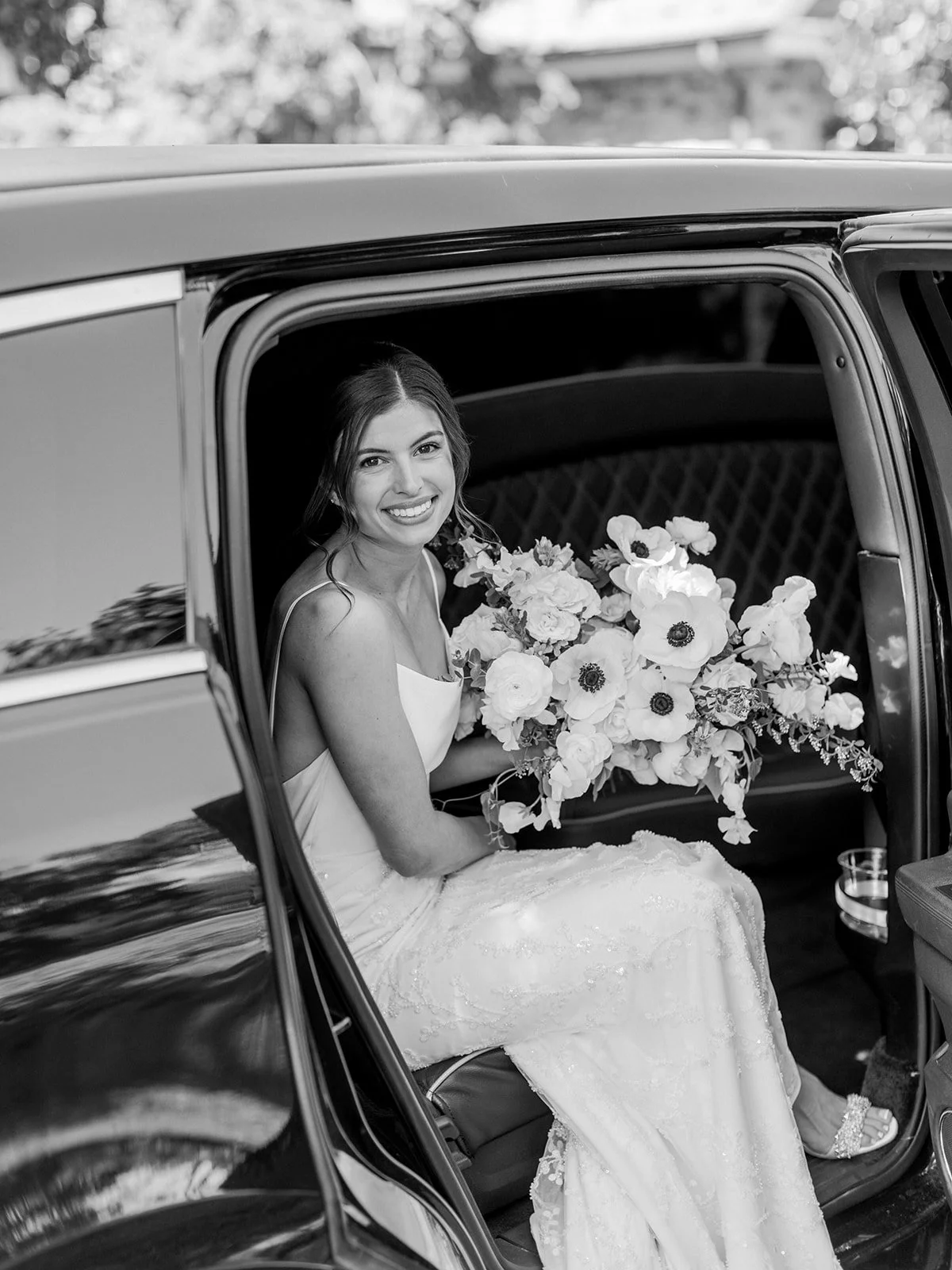 A smiling bride sitting inside a vintage car, holding a bouquet of flowers, wearing a wedding dress.