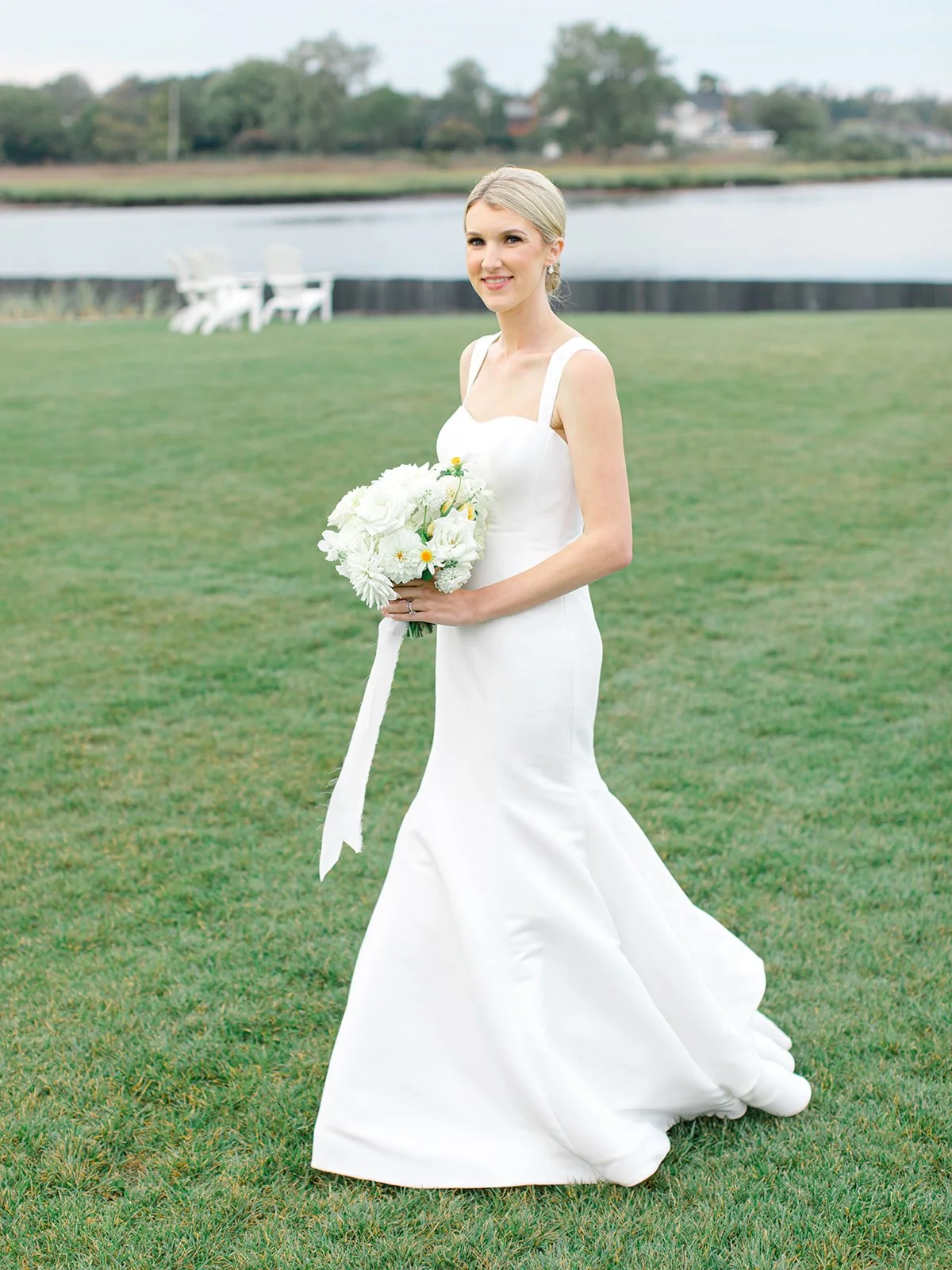 A bride in a white wedding gown holding a bouquet of white flowers, standing on a grassy area near a body of water with trees and chairs in the background.