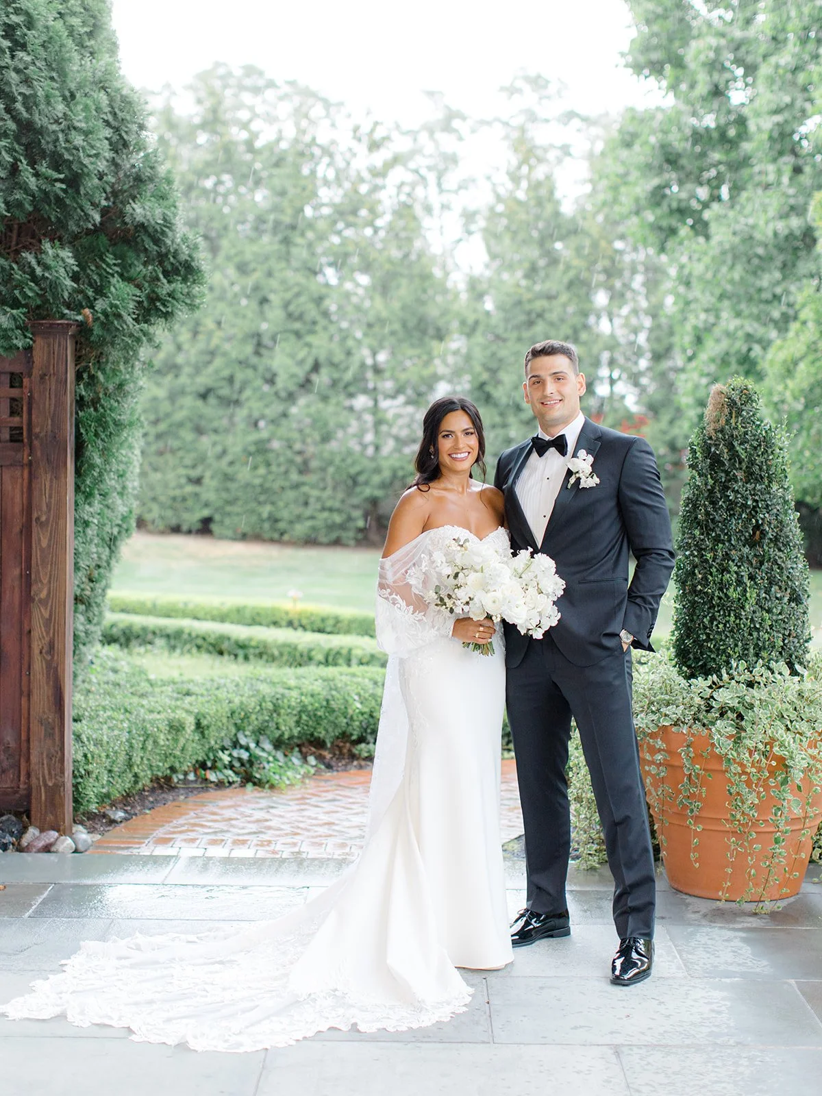 A bride and groom standing outdoors on a wedding day, smiling at the camera. The bride is wearing a white off-the-shoulder wedding gown and holding a bouquet of white flowers. The groom is dressed in a black tuxedo with a bow tie and boutonniere. The