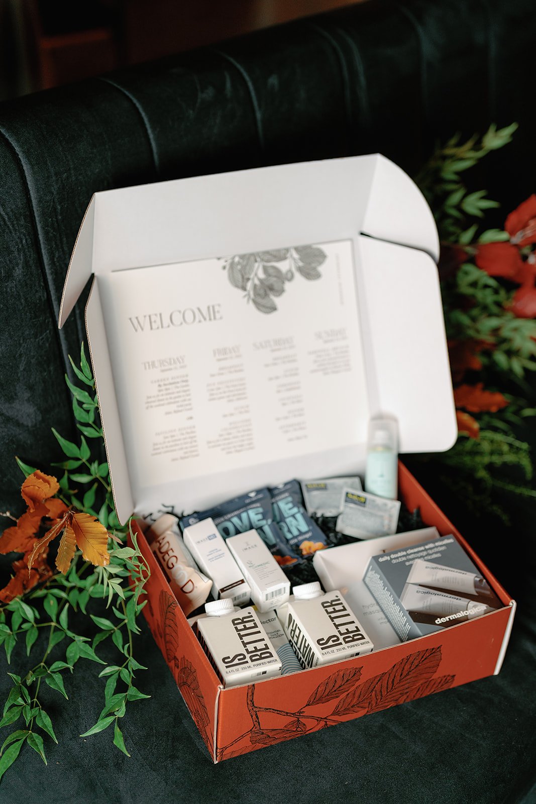 Open COVID-19 care package with various supplies, including lip balm, lotions, and medication, placed on a dark surface with decorative green and orange foliage around it.