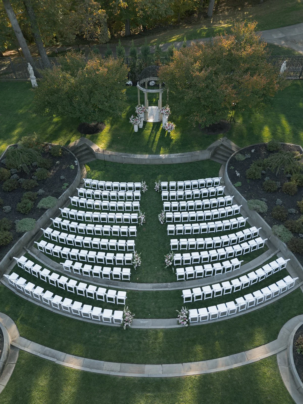 Empty wedding ceremony chairs arranged in rows with floral arrangements leading to a small gazebo, surrounded by landscaped garden and trees.