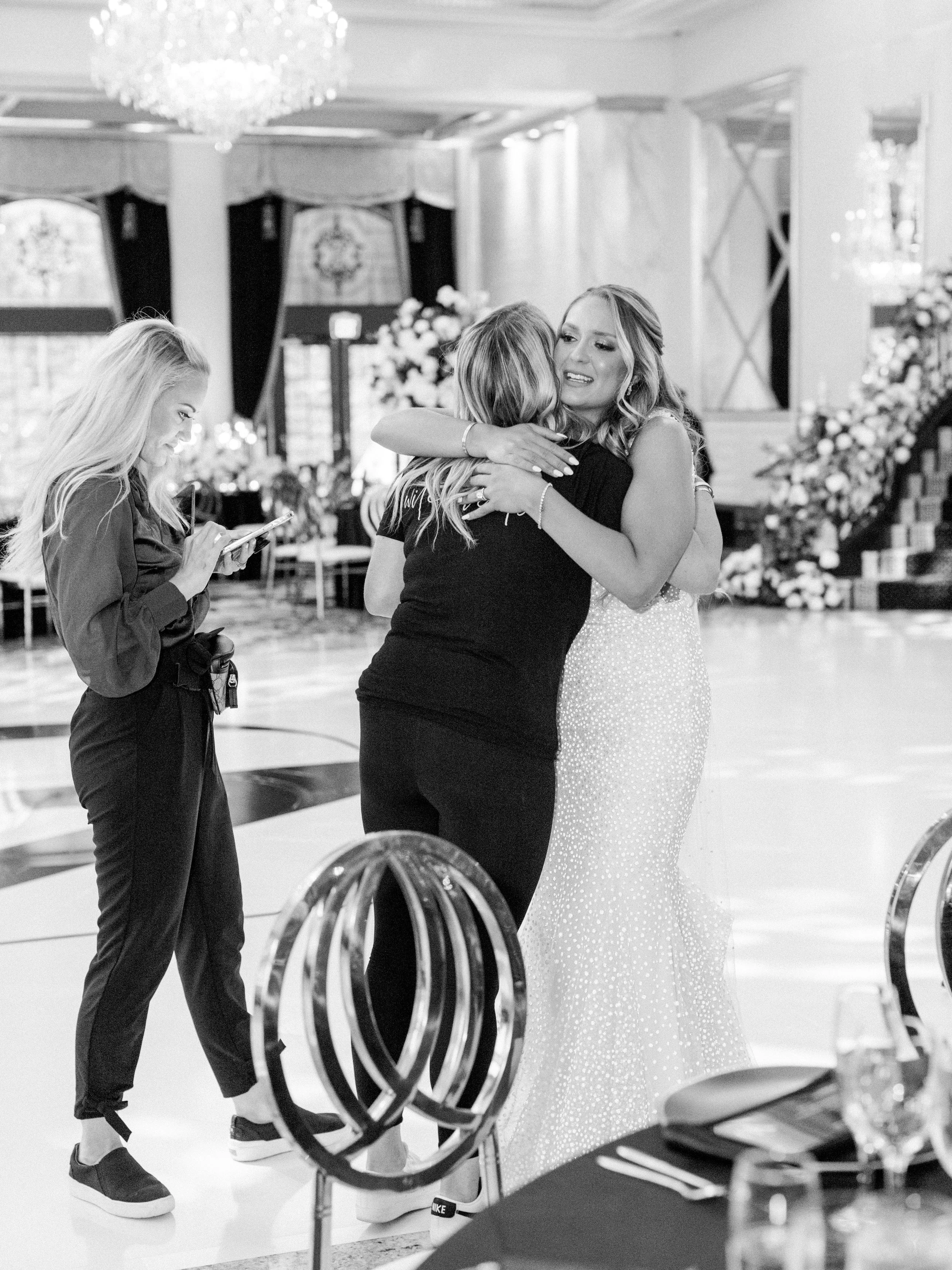 Two women hugging in a ballroom with floral decorations and a chandelier, one woman wearing a sparkling dress and the other in dark clothing, while another woman looks at her phone.