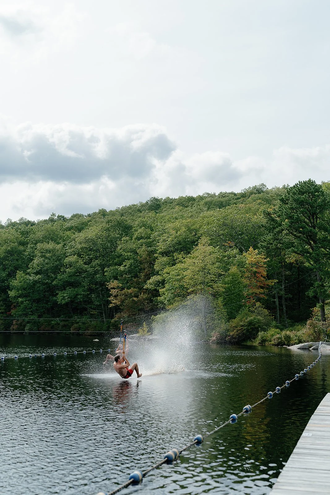 Person wakeboarding on a lake near a wooded shoreline with green trees, overcast sky, and a dock on the right side.