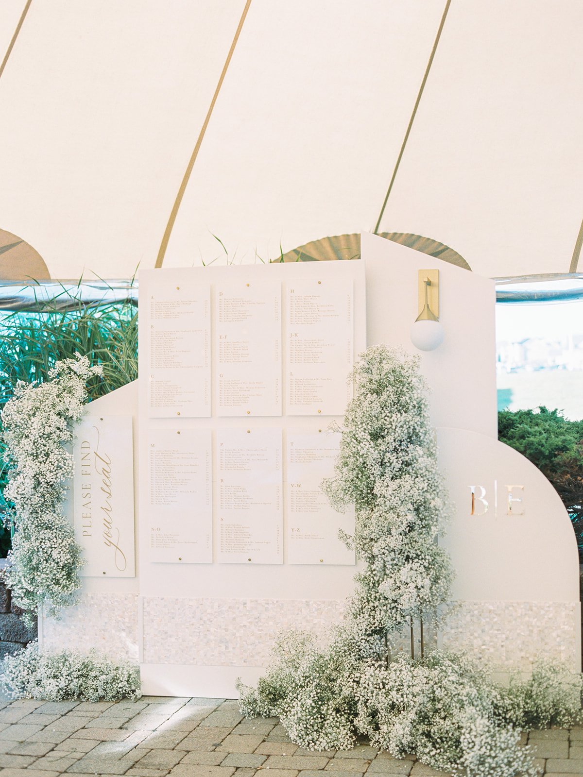 A seating chart display decorated with white flowers and greenery at a wedding or event, with a sign reading 'Please find your seat' on the left and 'BE' on the right, set against a white backdrop.