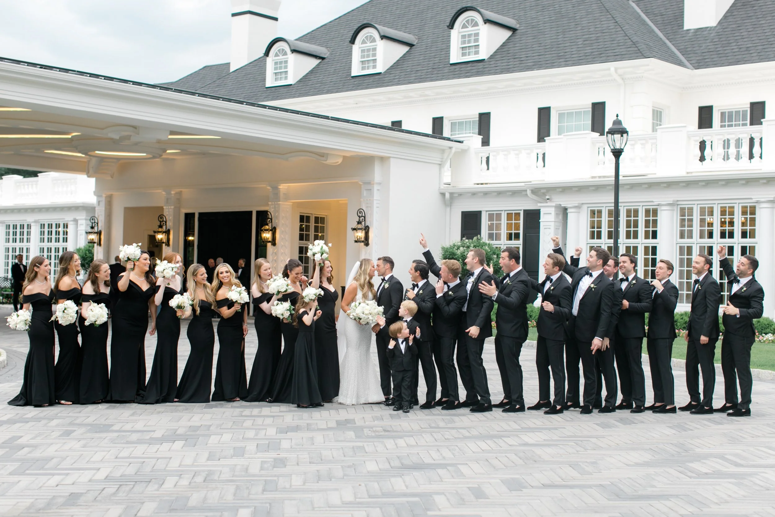 A wedding party standing outside a large white house with black shutters. The bride and groom are in the center, surrounded by bridesmaids holding bouquets and groomsmen. They are celebrating, some with raised fists and smiling.