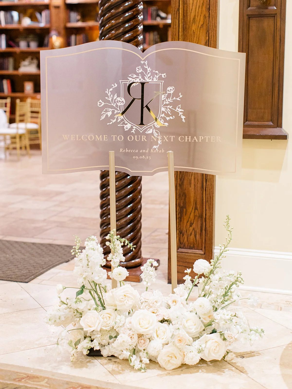 A welcoming sign at a wedding or event, featuring a large open book design with initials and text, surrounded by white flowers on the floor.