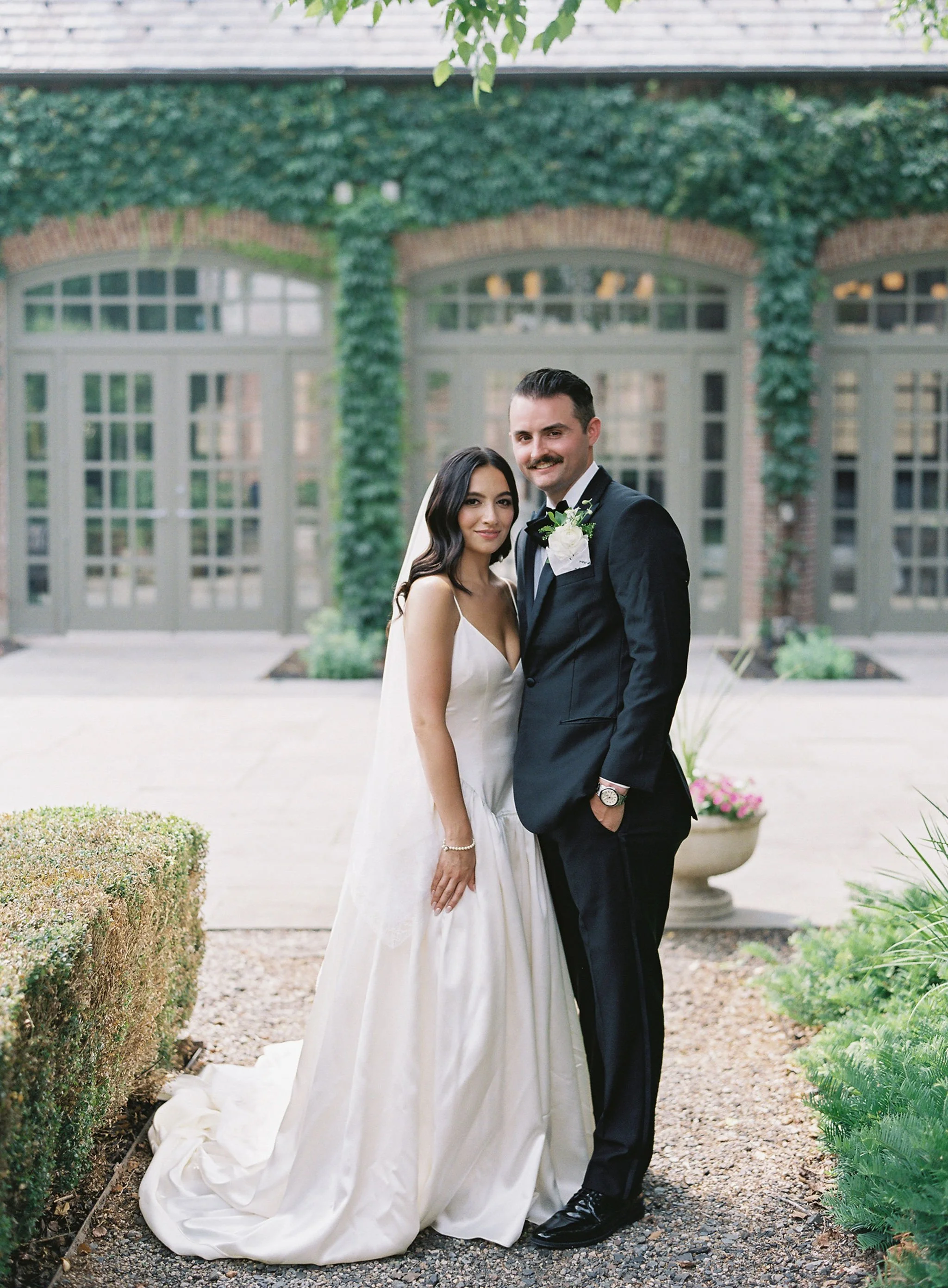 A bride and groom standing together outdoors, in front of a building with green ivy and large windows, during their wedding.