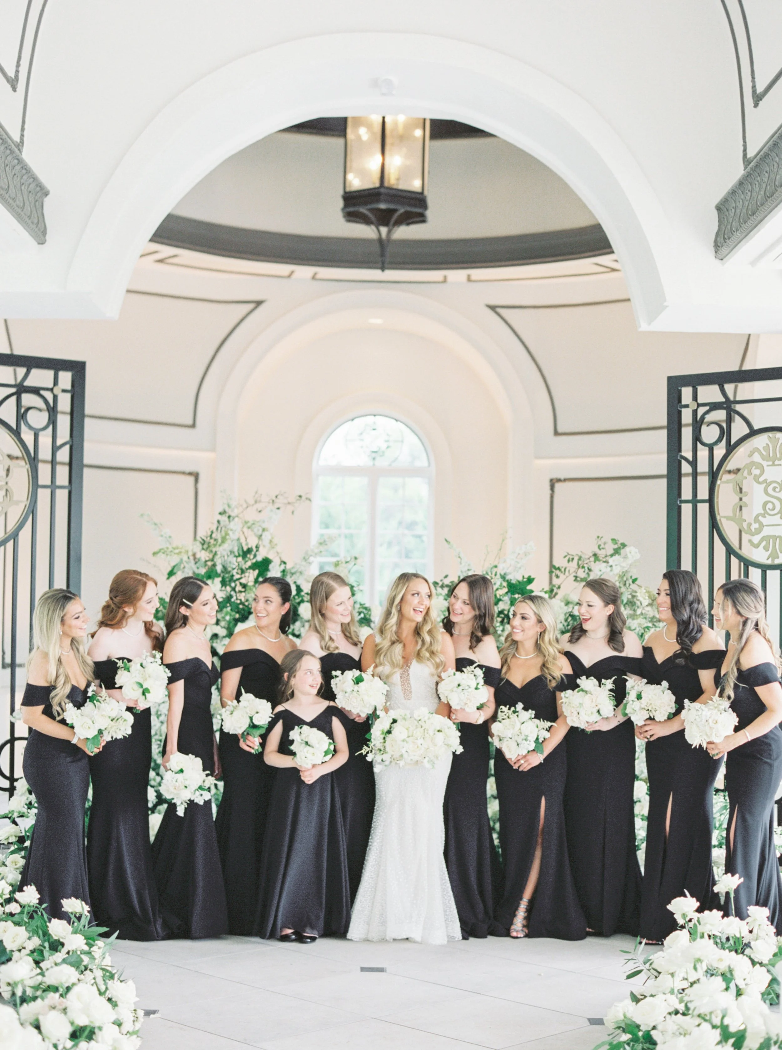 A bride in a white wedding dress surrounded by bridesmaids in black dresses, all holding white bouquets, inside a decorated indoor venue with white flowers and greenery.