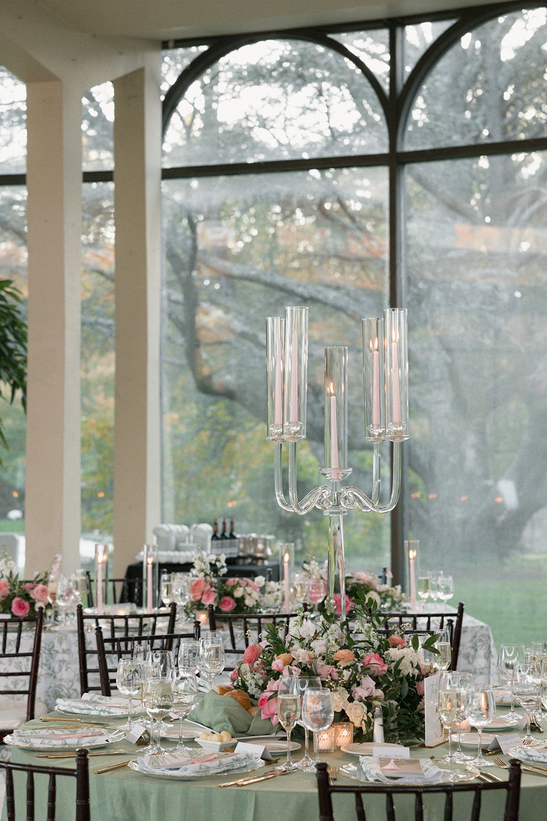 Elegant banquet table decorated with floral arrangements and a tall glass candelabra, set inside a venue with large windows overlooking trees.