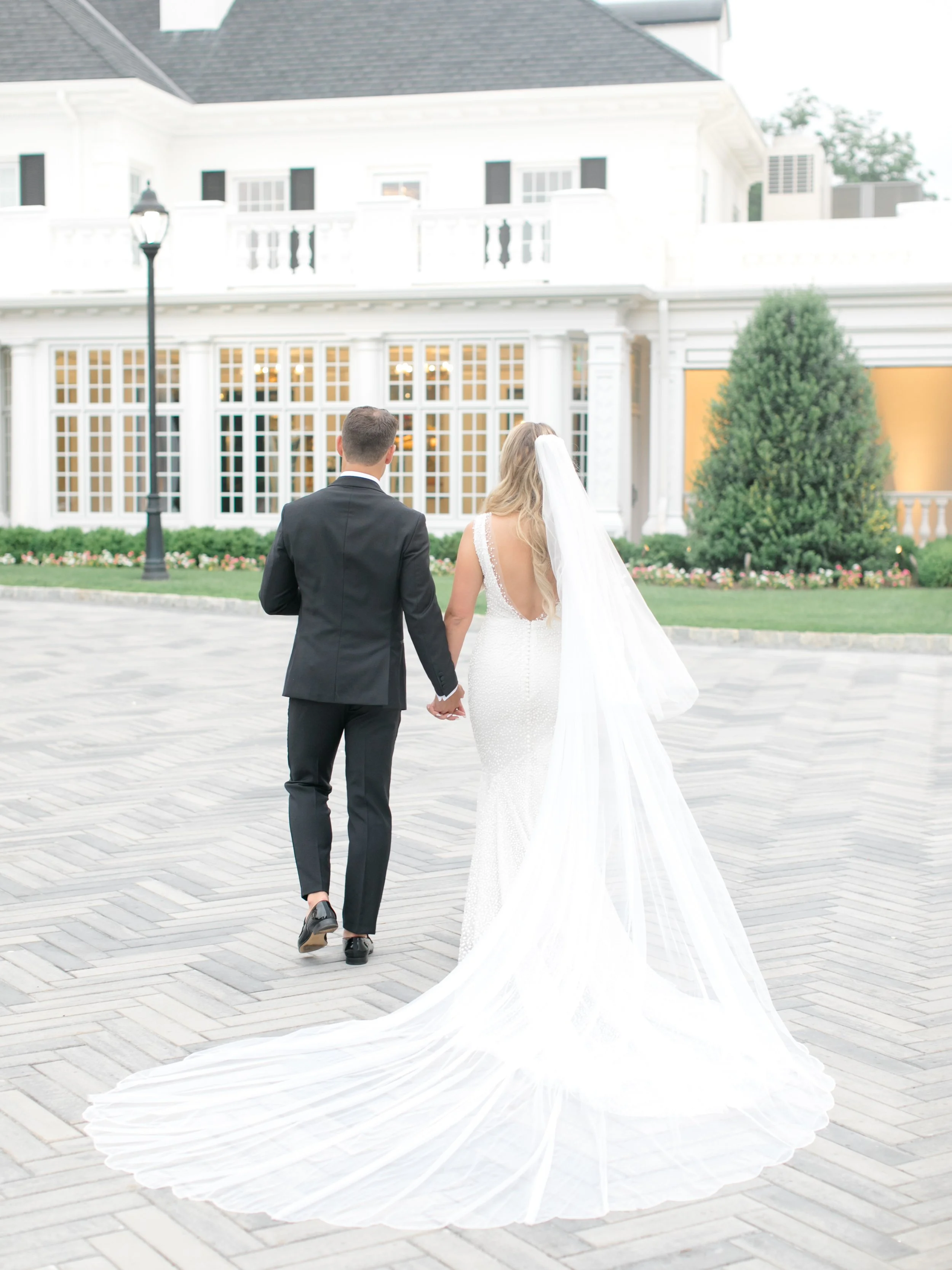 A bride and groom walking hand in hand outside a white building with large windows and a manicured lawn.