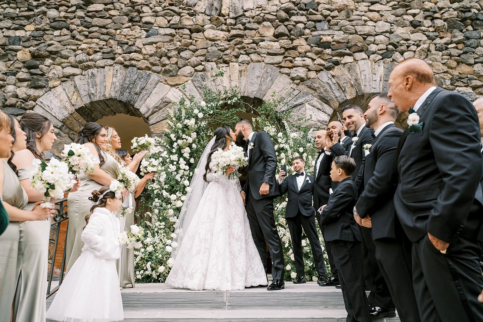 A wedding ceremony with a bride and groom sharing a kiss in front of a stone archway decorated with white flowers, surrounded by wedding guests in formal attire.