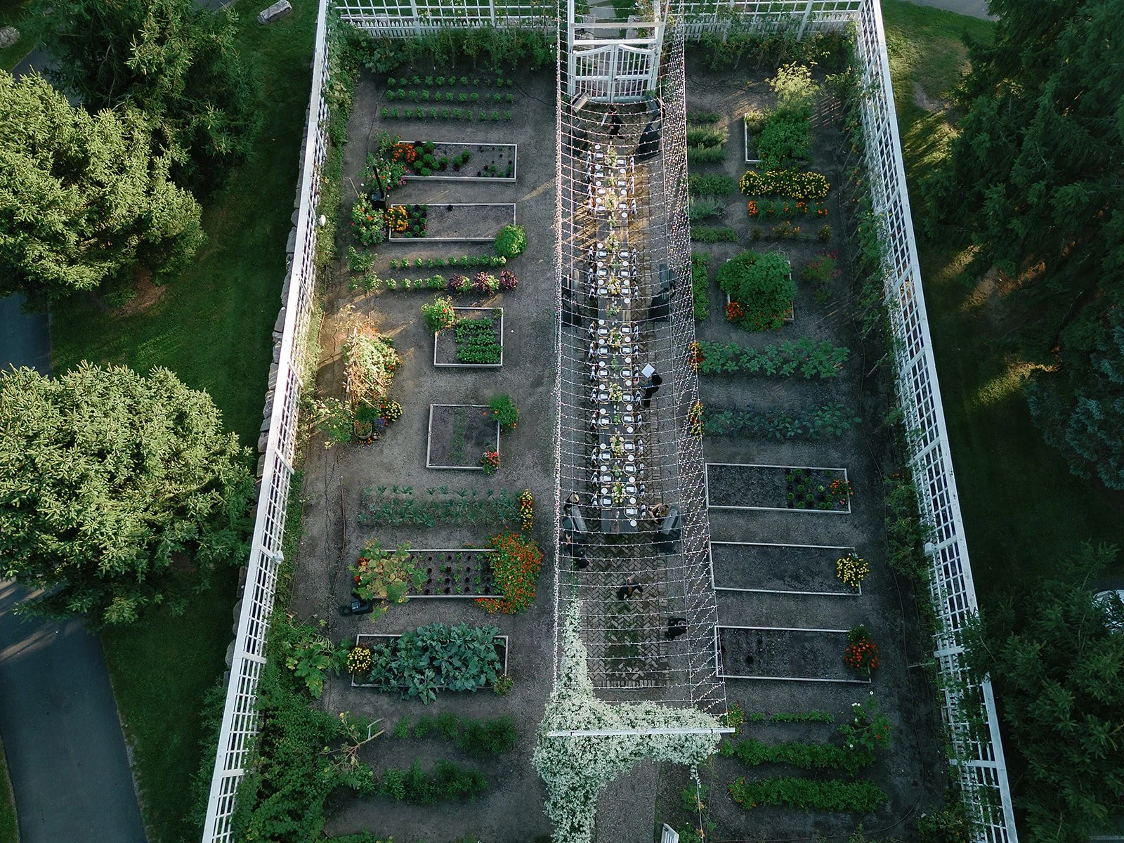 An aerial view of a large garden with various flower beds and vegetable plots, surrounded by a white fence. A long table set for an outdoor event runs down the center, with chairs around it. People are gathered around the table, and a white floral ar
