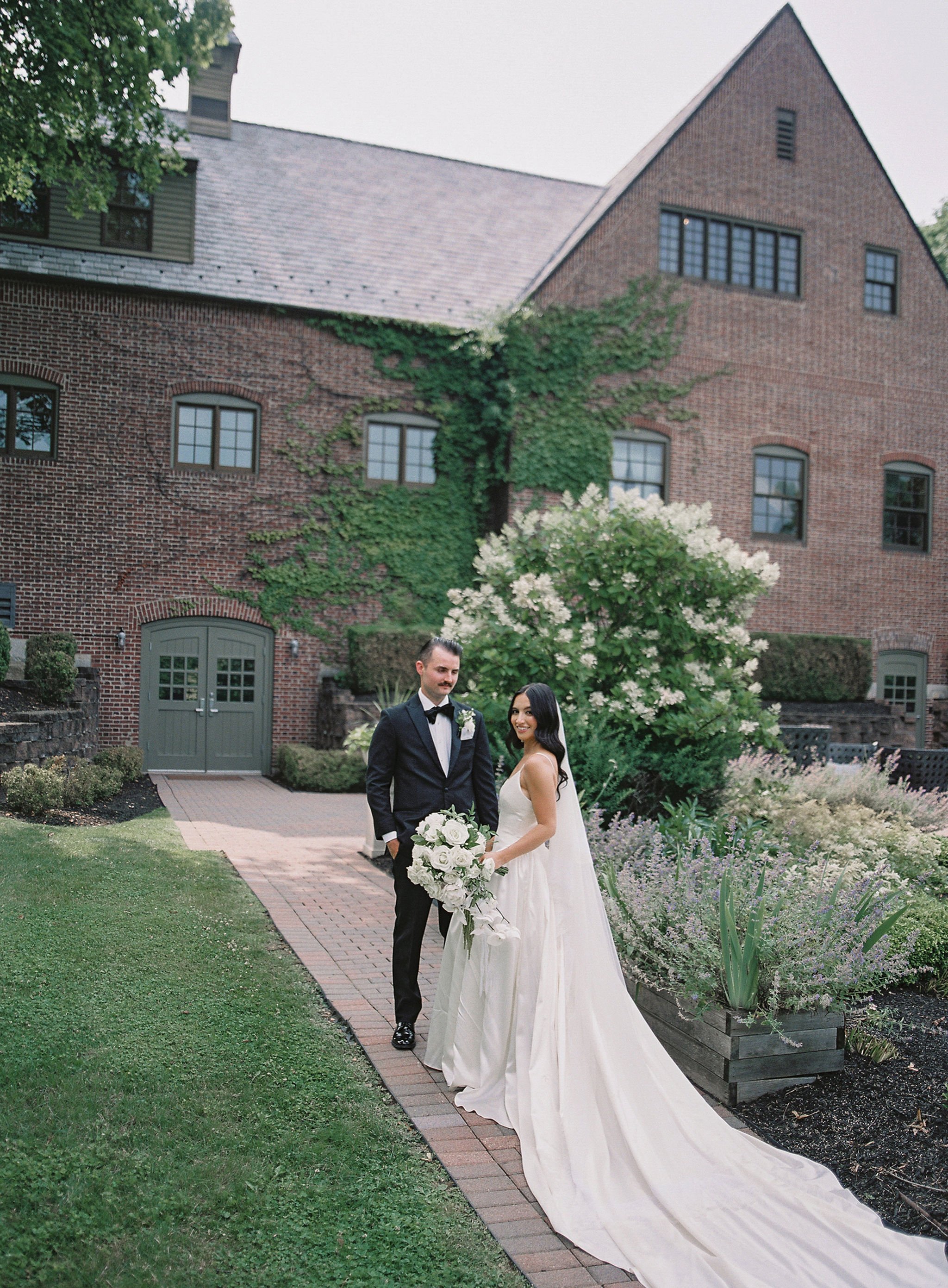 A bride in a white wedding gown holding a bouquet and a groom in a tuxedo standing on a garden pathway in front of a brick mansion with greenery and flowering bushes.