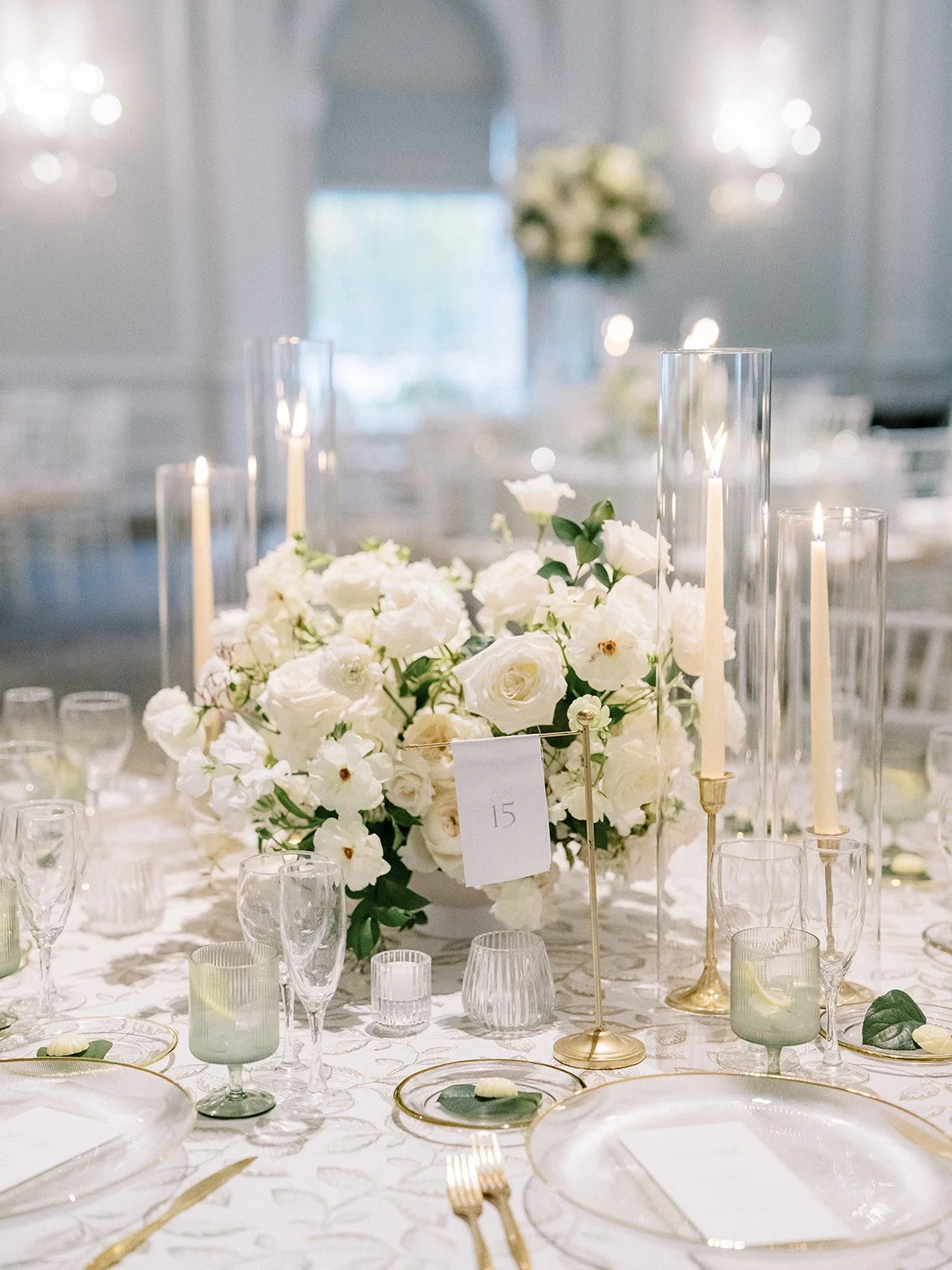 Elegant banquet table decorated with a large white floral centerpiece, tall glass candle holders with lit taper candles, and glassware set for a formal event in a bright room.