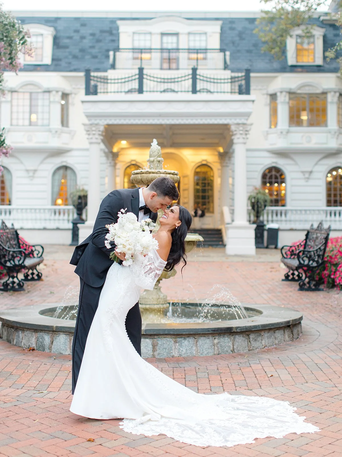 A newlywed couple smiling and laughing in a romantic pose in front of a historic mansion with a fountain, brick walkway, and garden flowers, during sunset.