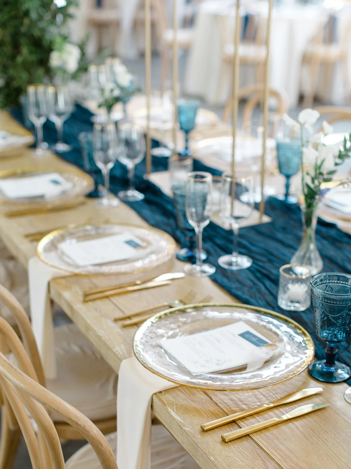 Table set for a formal event with gold-rimmed transparent plates, gold cutlery, blue glassware, and white napkins with menus, decorated with white flowers and greenery.