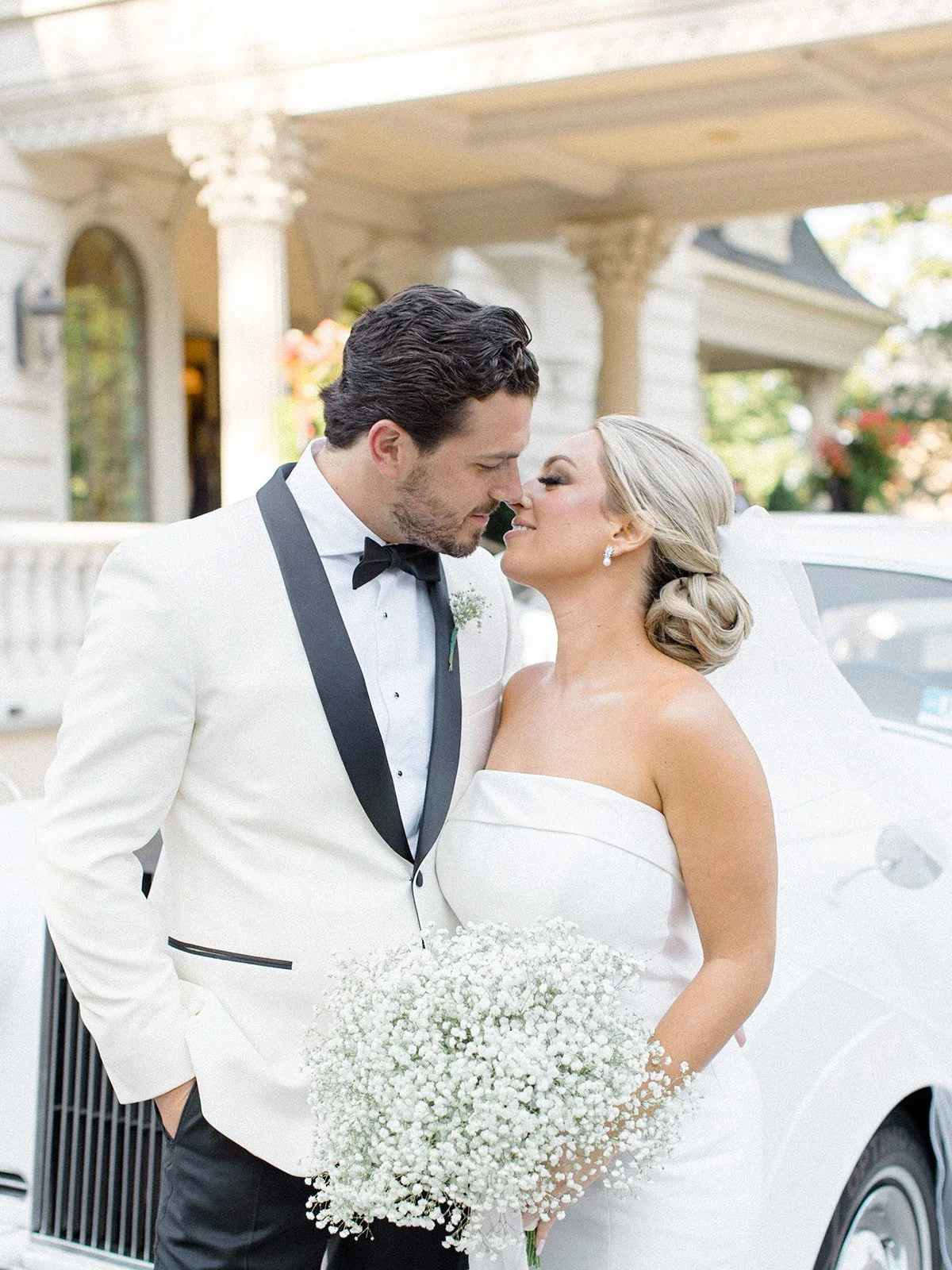 A bride and groom stand close together, about to kiss, outdoors in front of a white luxury car, with a large white house in the background.