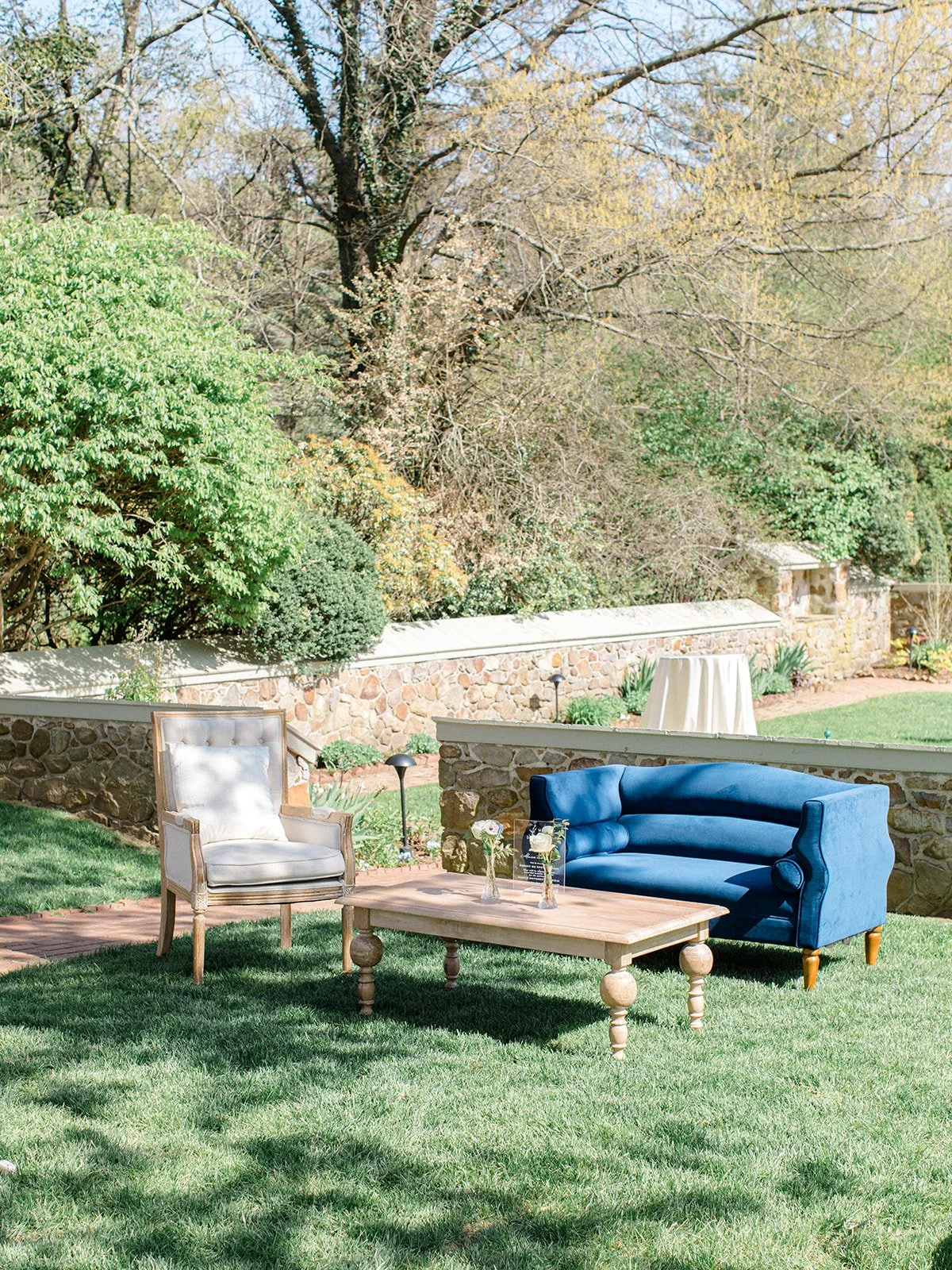 An outdoor patio with a vintage armchair, a blue sofa, and a wooden coffee table with vases, surrounded by green grass and trees.