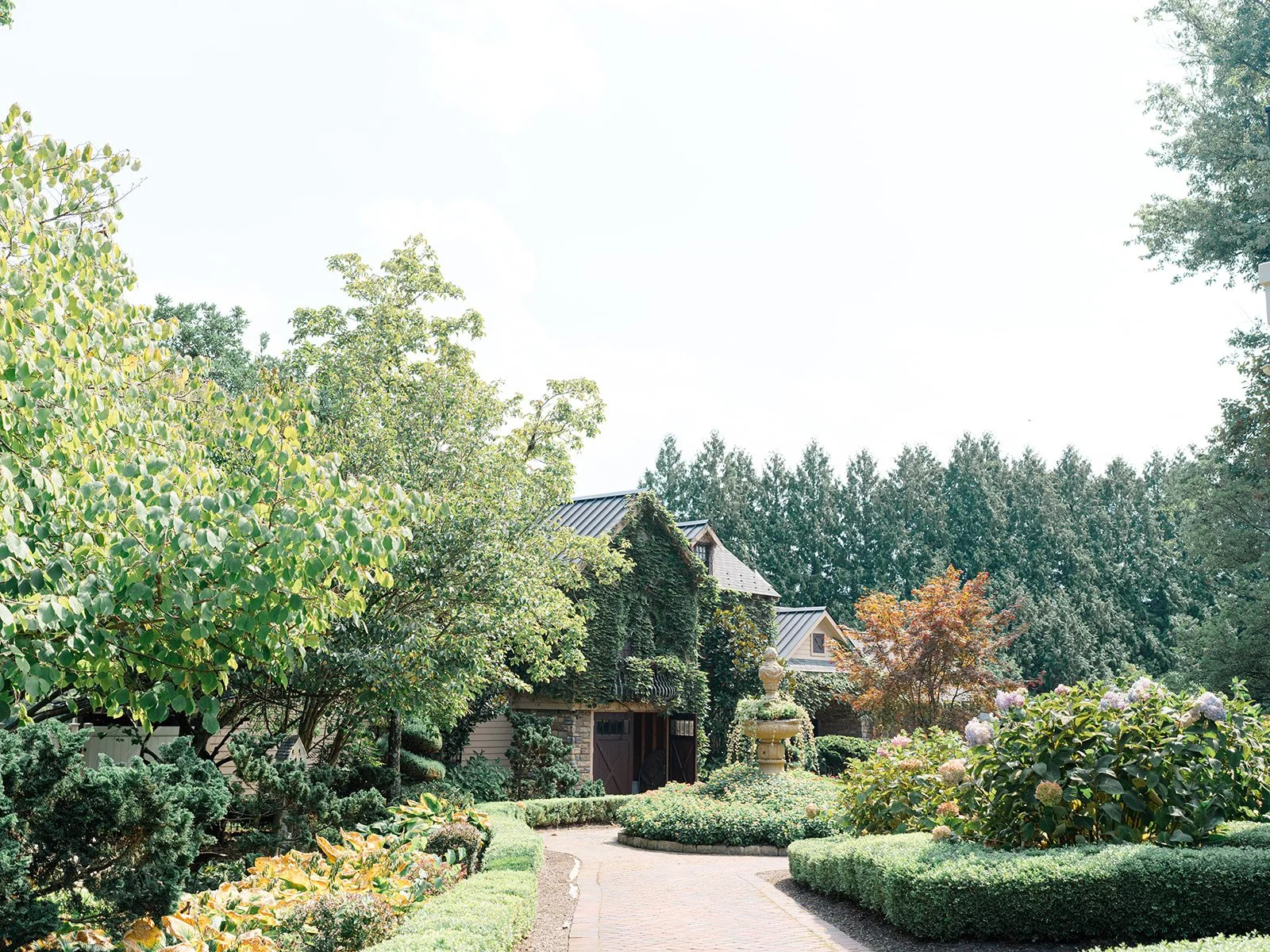 A landscaped garden with a brick pathway, lush green trees and bushes, and a stone fountain in front of a house with ivy-covered walls and a sharp roof.