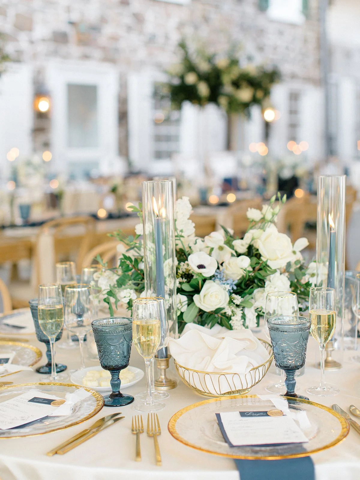 Elegant banquet table set with gold-rimmed plates, gold utensils, champagne glasses, dark blue wine glasses, tall candle holders, and a large floral centerpiece with white and blue flowers. The background shows a decorated indoor venue with white wal