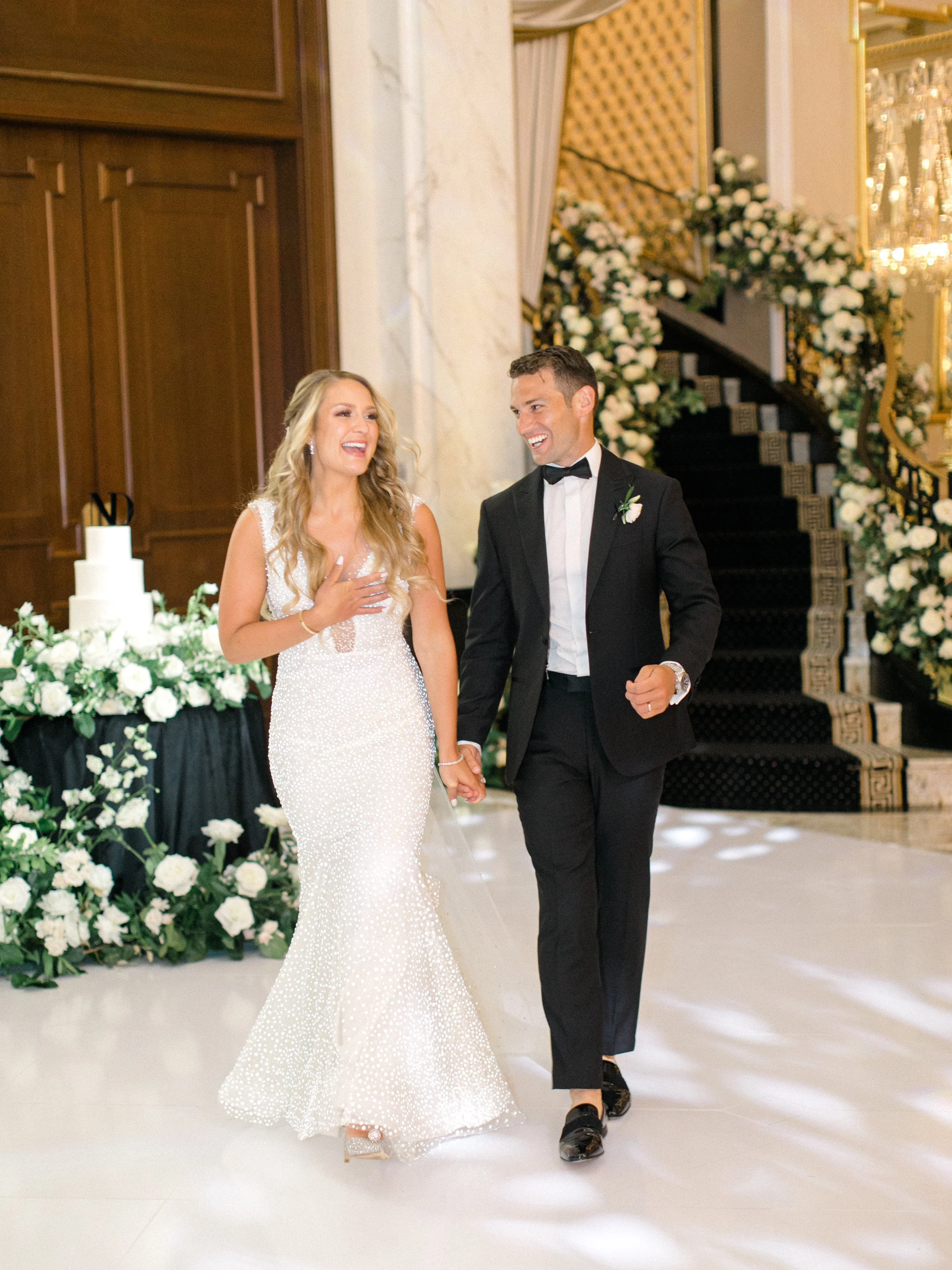 A bride and groom walking hand in hand during their wedding celebration, smiling and sharing a joyful moment indoors decorated with white flowers and elegant decor.