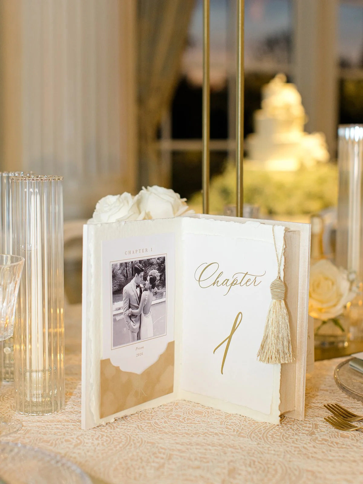 Decorative wedding table centerpiece featuring an open book with a black-and-white wedding photo on the left page and the word "Chapter" with a gold tassel on the right page, surrounded by white roses and candles.