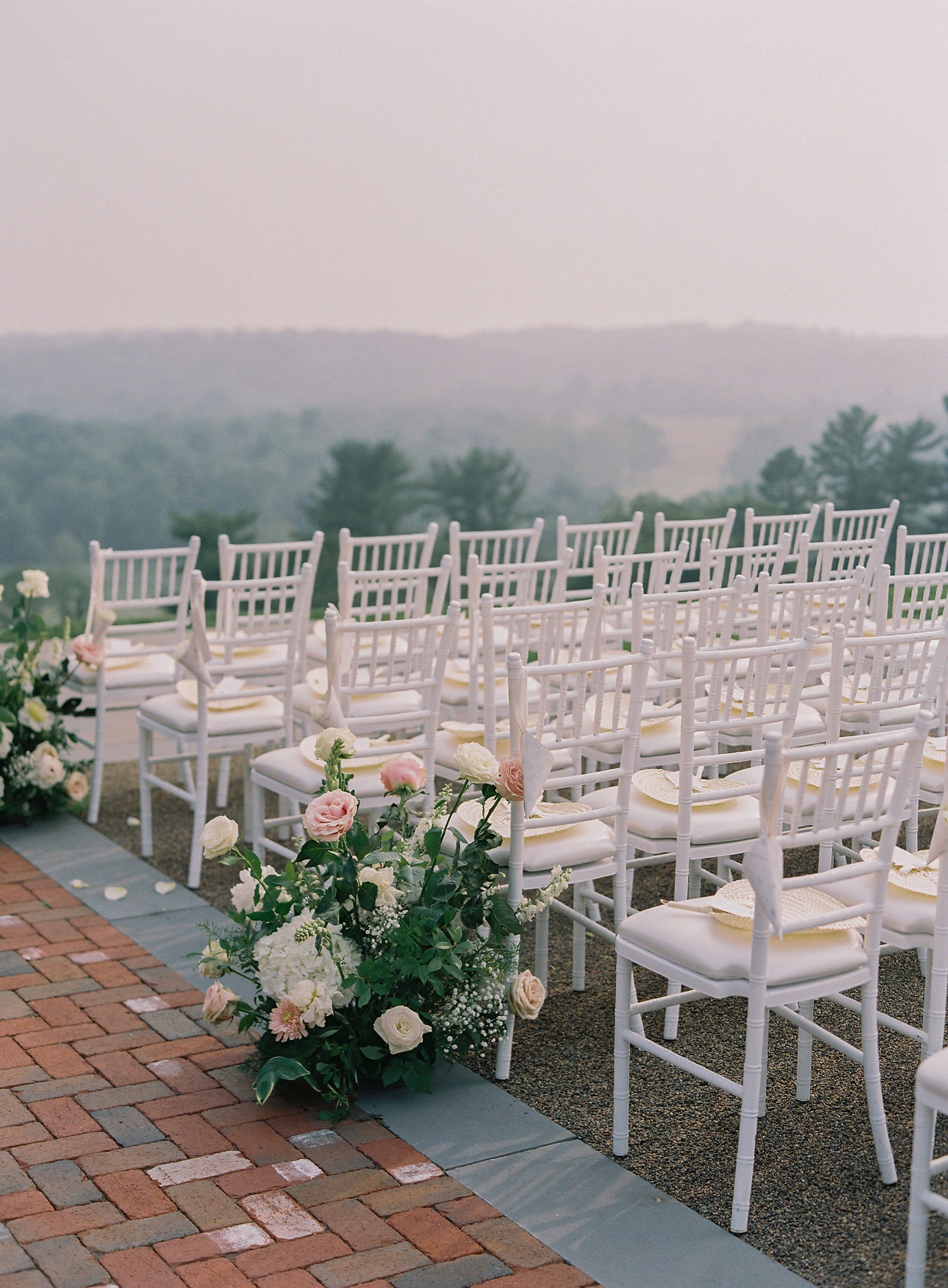Outdoor wedding setup with white chairs, floral arrangements, and a scenic background.