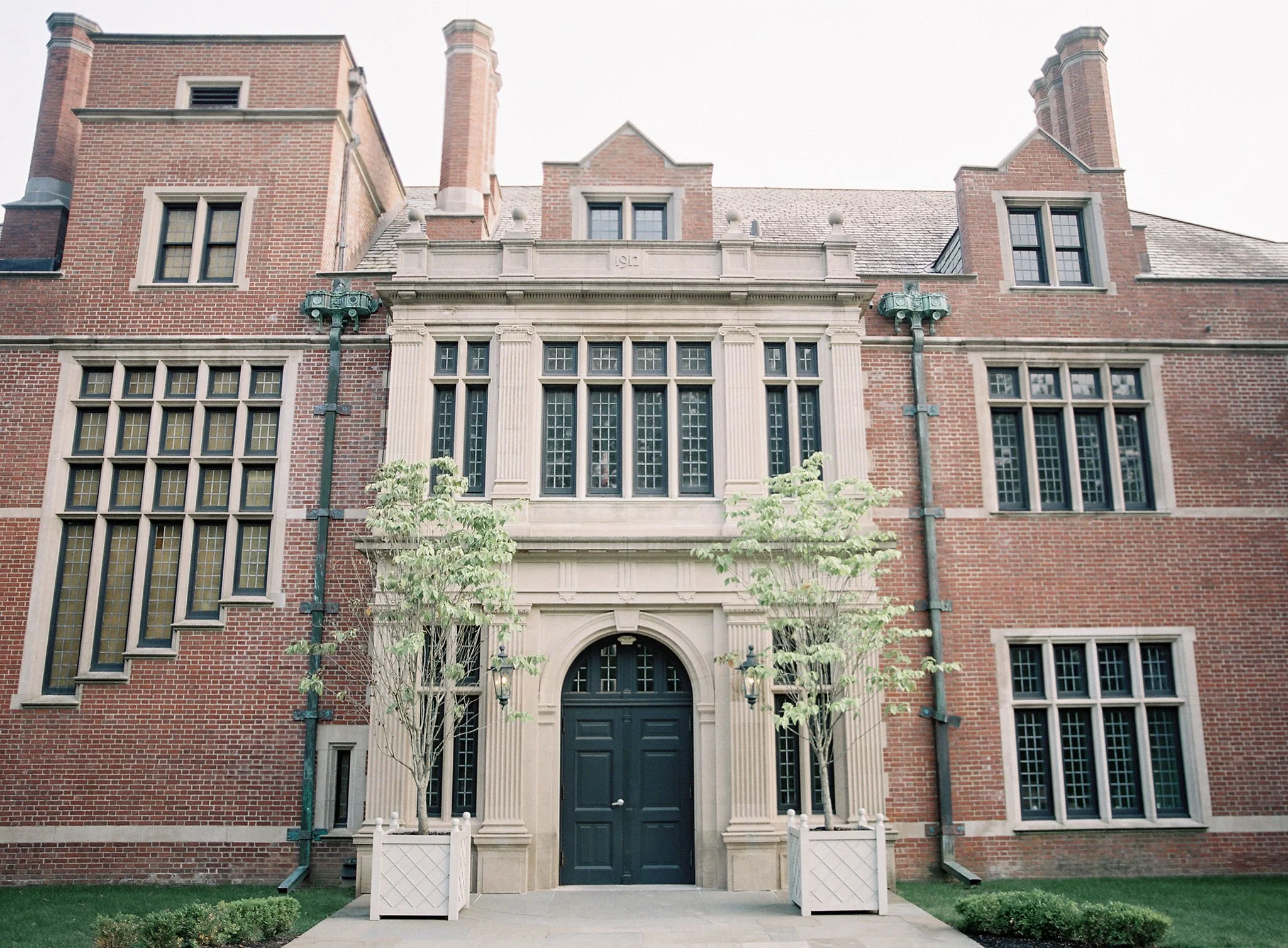 Front view of a historic brick building with large windows, an arched black door, decorative stone columns, and two small trees in white planters flanking the entrance.