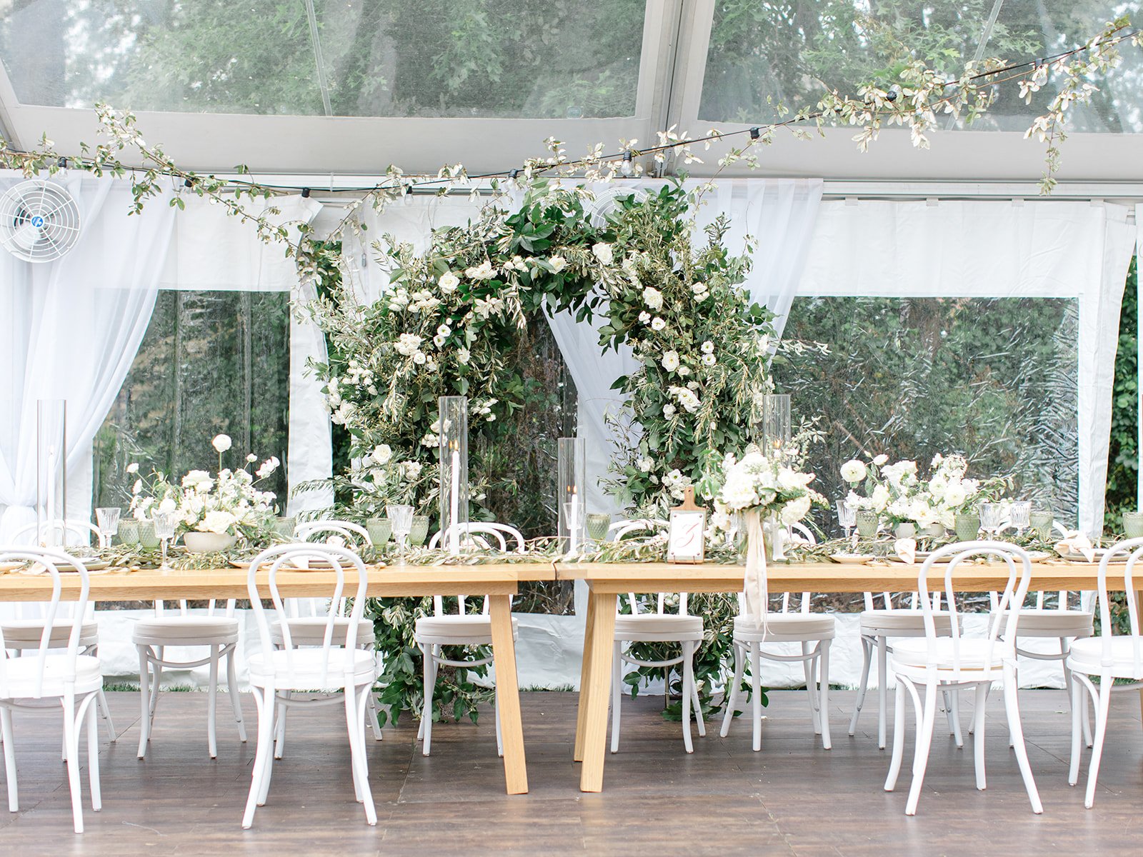 Buffet or dining table setup with white chairs, floral arrangements, candles, and greenery in a decorated outdoor or tented space with white curtains and a large floral arch in the background.