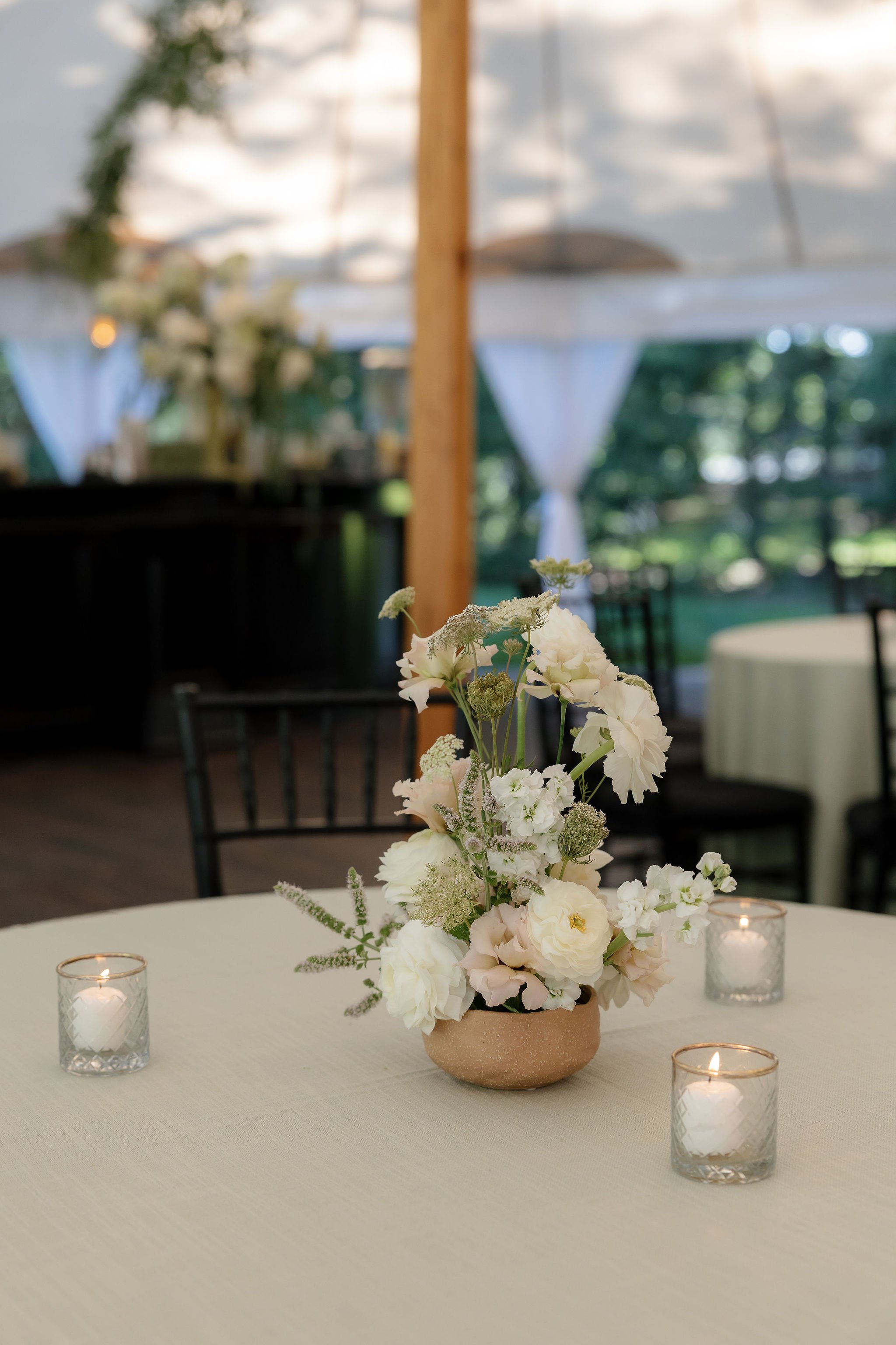 A white floral centerpiece on an elegant event table, surrounded by three lit candles in glass holders, with a blurred outdoor backdrop.