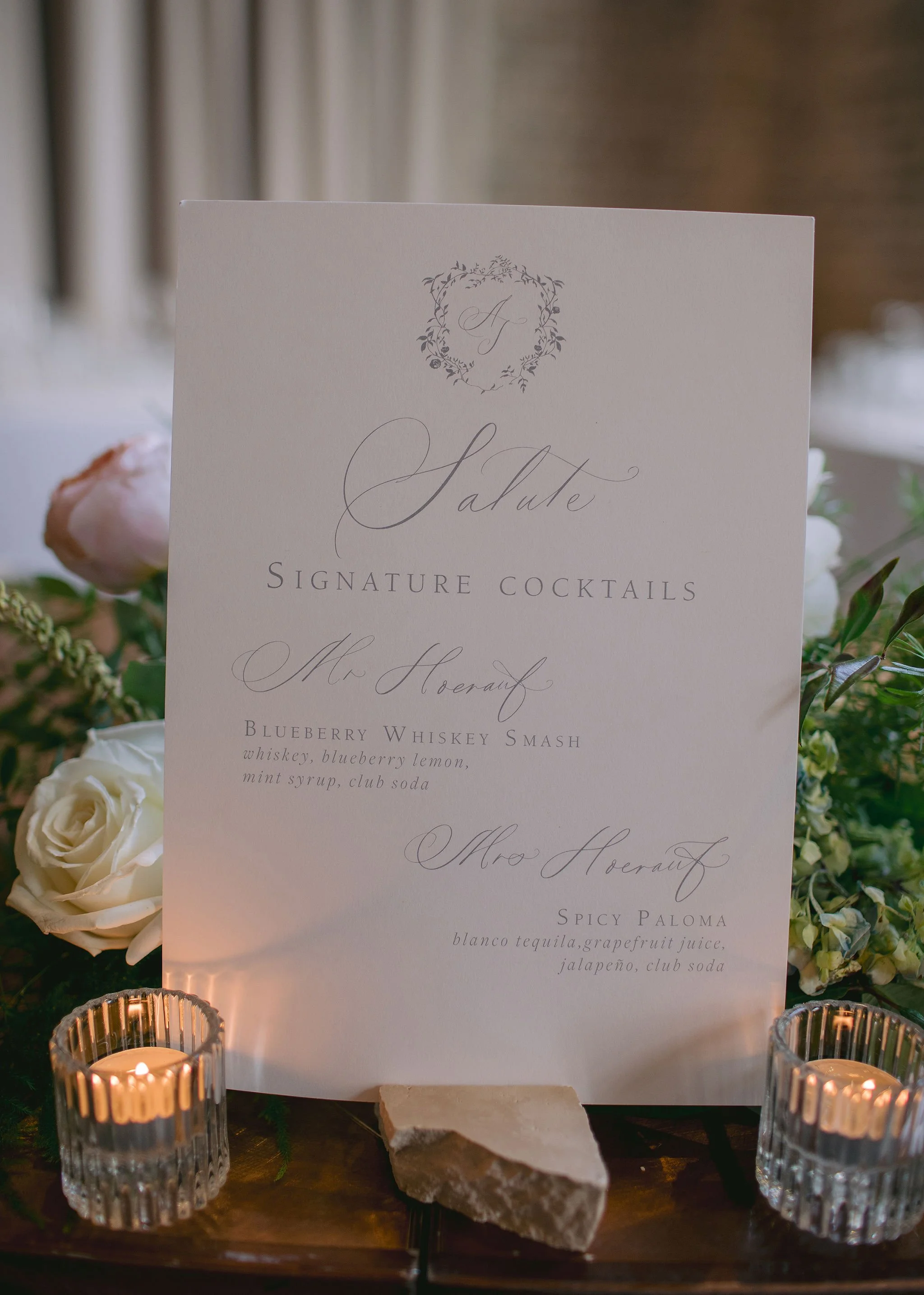 Wedding cocktail menu featuring signature drinks, displayed on a table with flowers, candles, and a small stone, with elegant cursive printing.