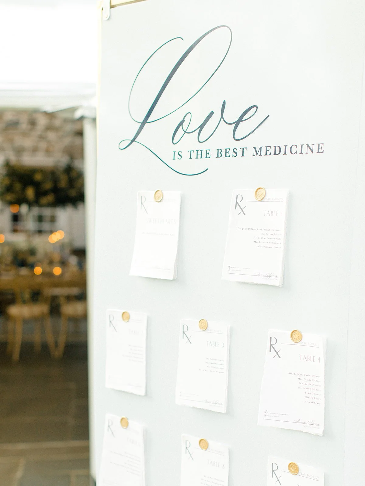 A white board with the phrase 'Love is the best medicine' written at the top in elegant cursive. Below, there are paper notices pinned with gold seals, each mentioning different tables and names, possibly a seating chart or event schedule.
