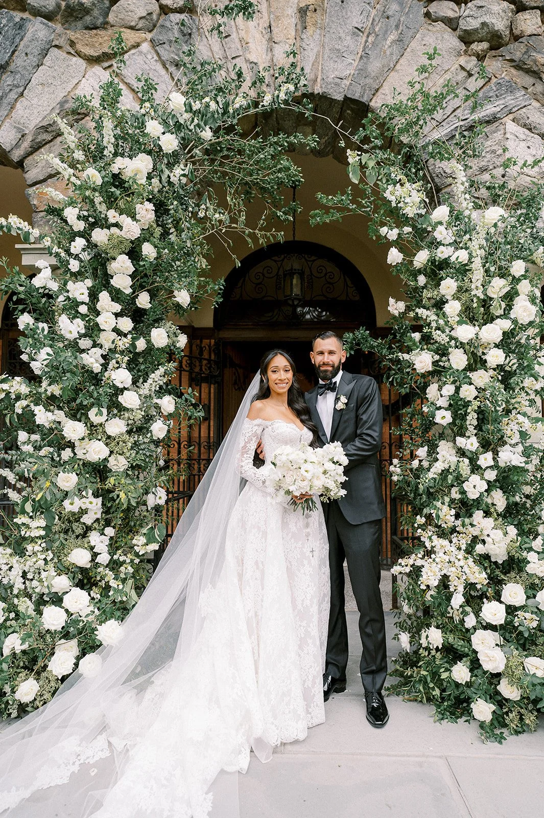 A bride and groom standing under a large floral arch of white roses and greenery, smiling at the camera. The bride is in a lace wedding gown with a long veil, holding a bouquet of white flowers. The groom is in a black tuxedo with a bow tie, standing