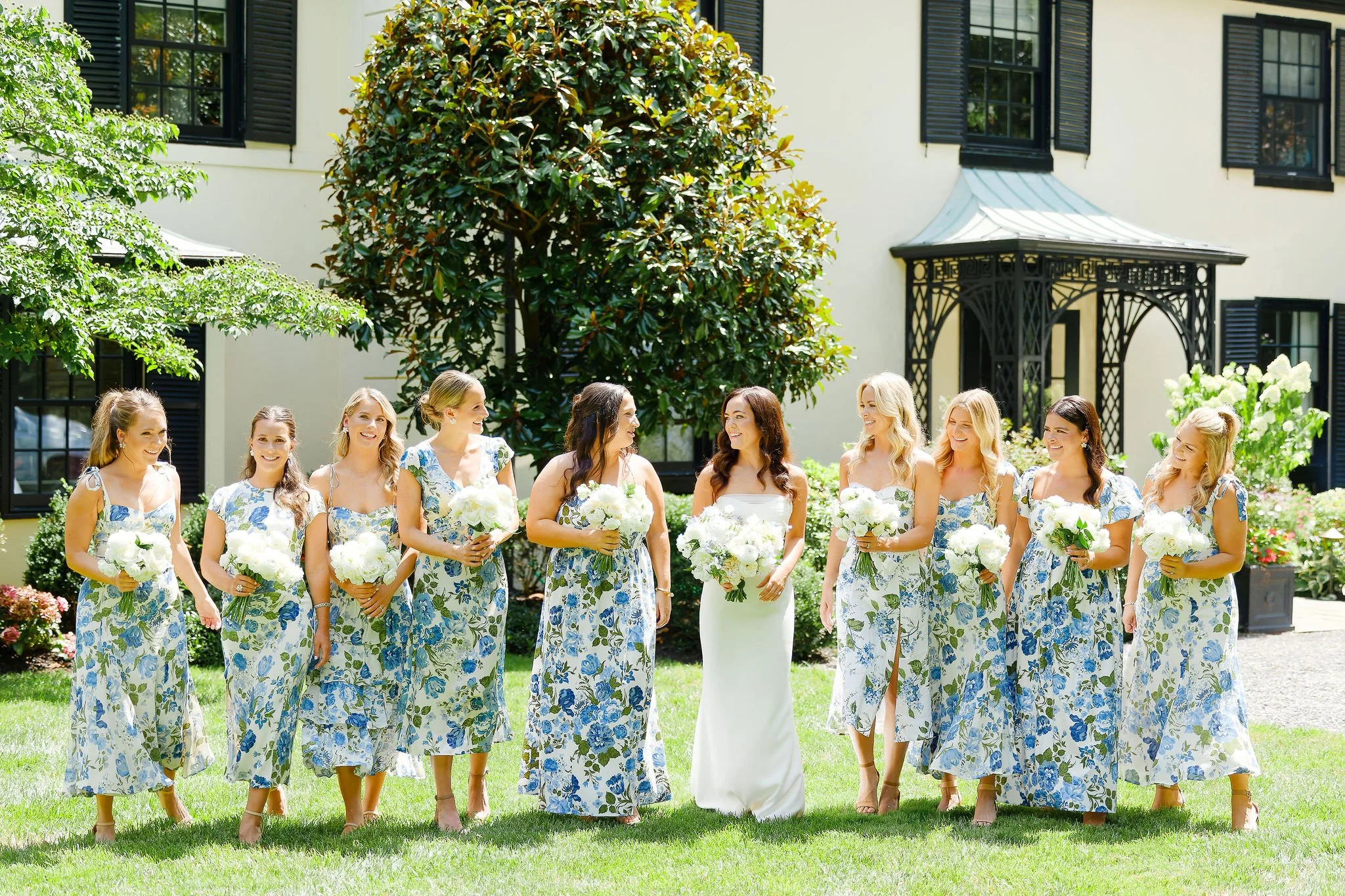 A bride and her nine bridesmaids standing outdoors on a sunny day in front of a yellow house with black shutters, holding white bouquets, all dressed in matching blue and white floral dresses.