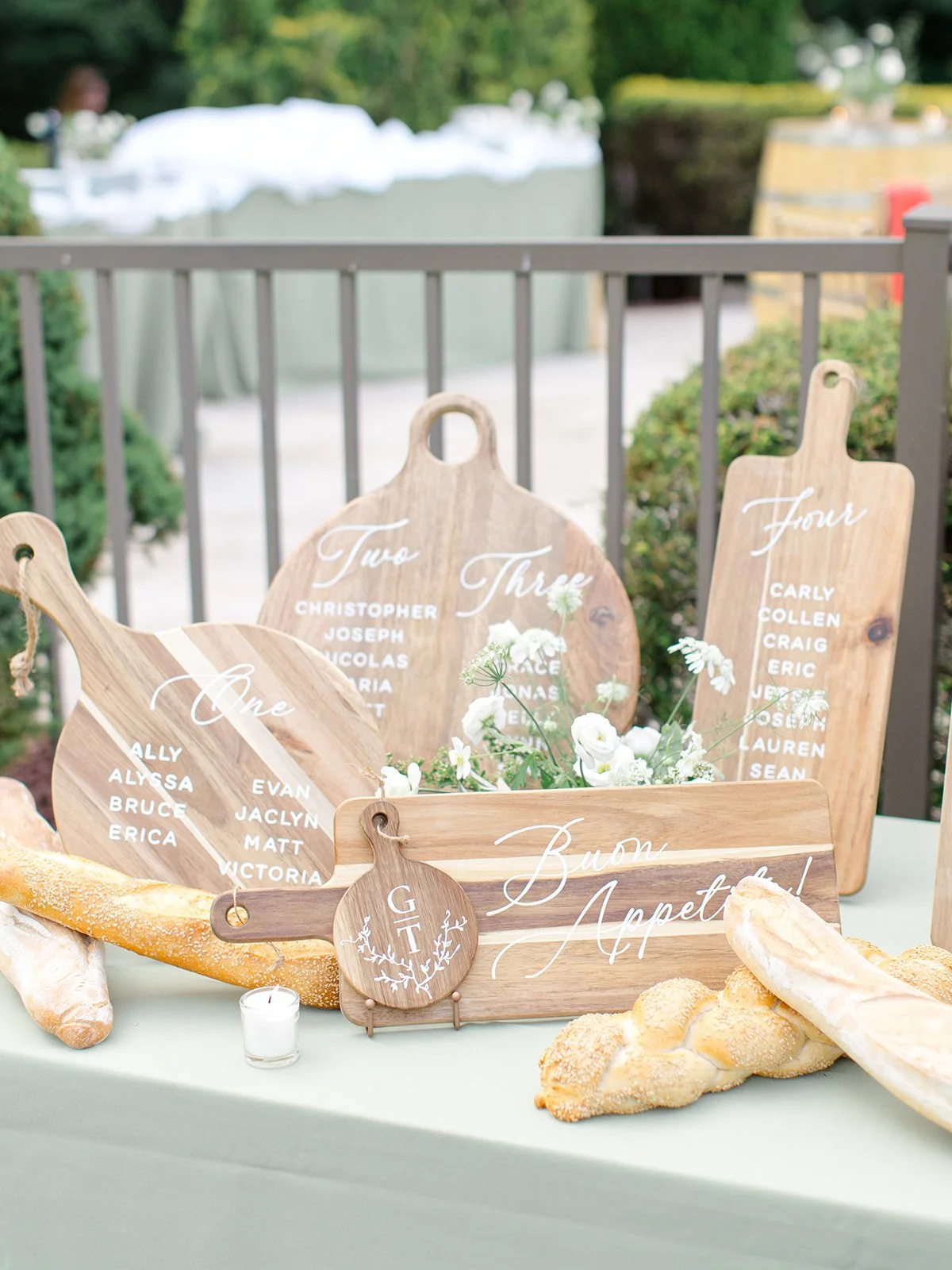 Wedding table place cards with names and table numbers, a sign saying 'Buon Appetito,' bread, flowers, and a small candle, set outdoors.
