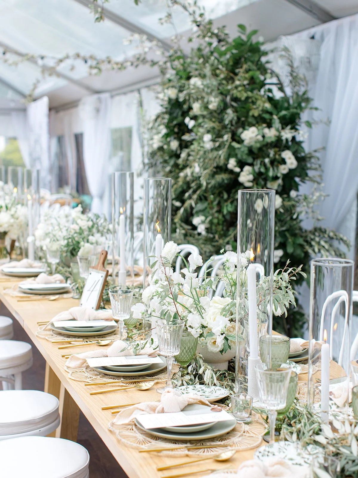 A decorated wedding or event table with white floral arrangements, tall glass candle holders, and matching tableware in a bright tented setting.