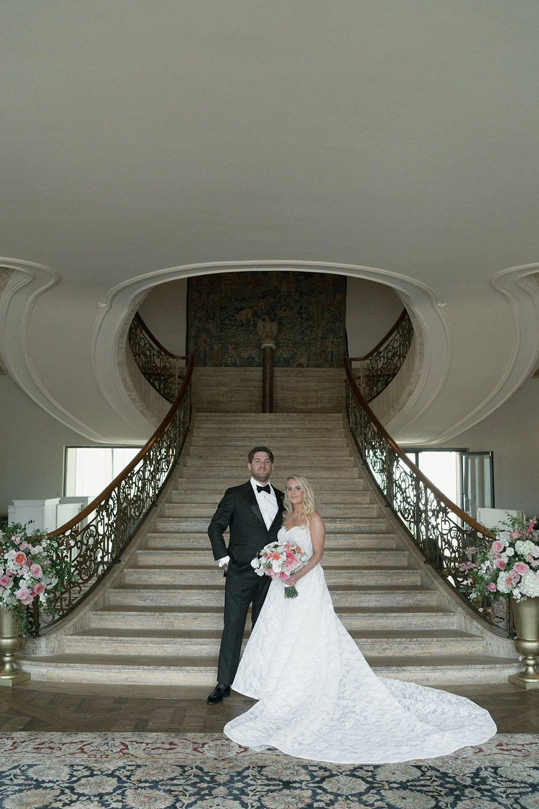 A bride and groom standing at the bottom of a grand staircase in an elegant venue, with bride holding a bouquet of flowers and wearing a white wedding gown, groom in a black tuxedo.