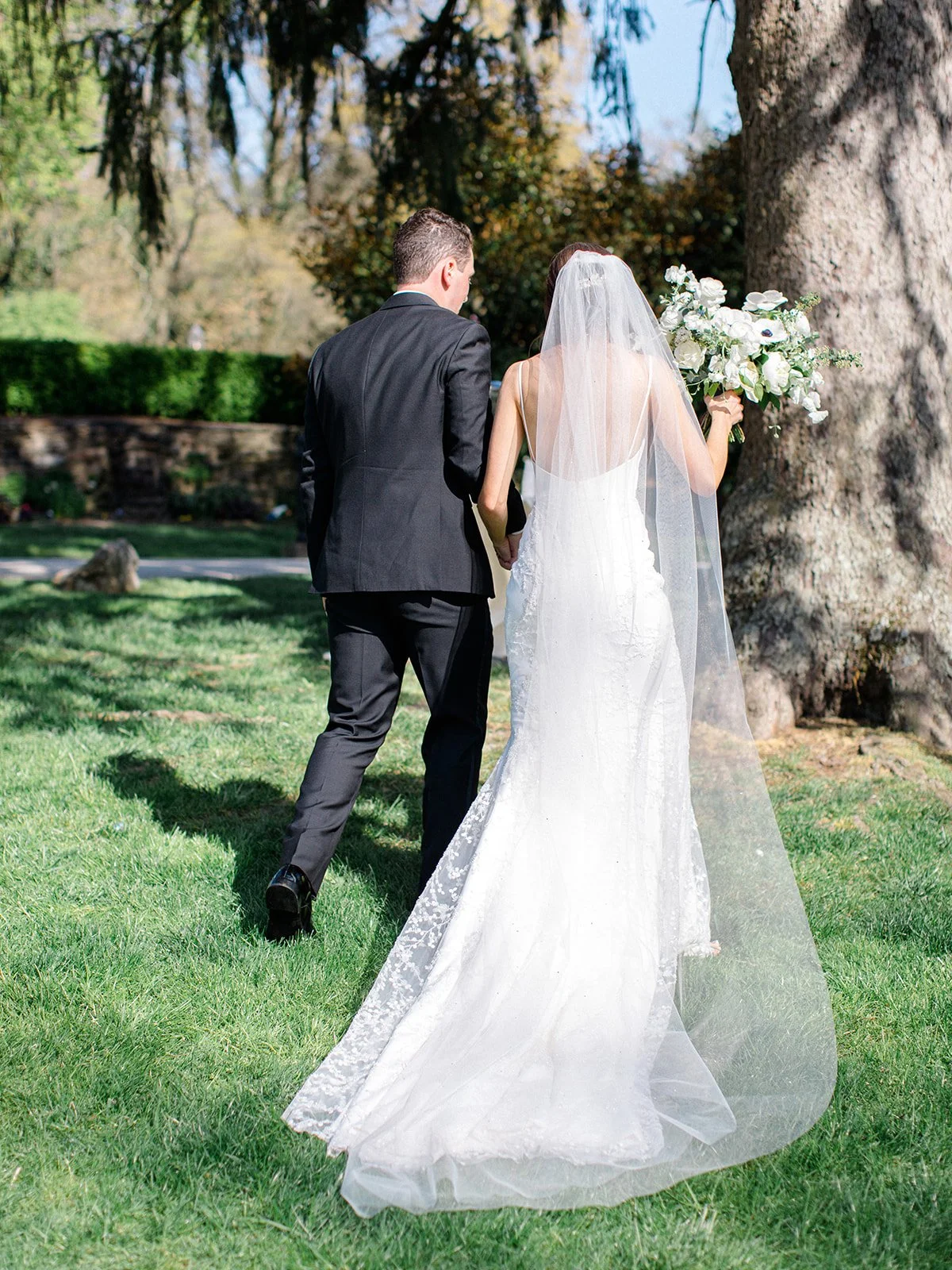 Bride in a white wedding dress and veil holding a bouquet walking with a groom in a black suit outdoors near a large tree.