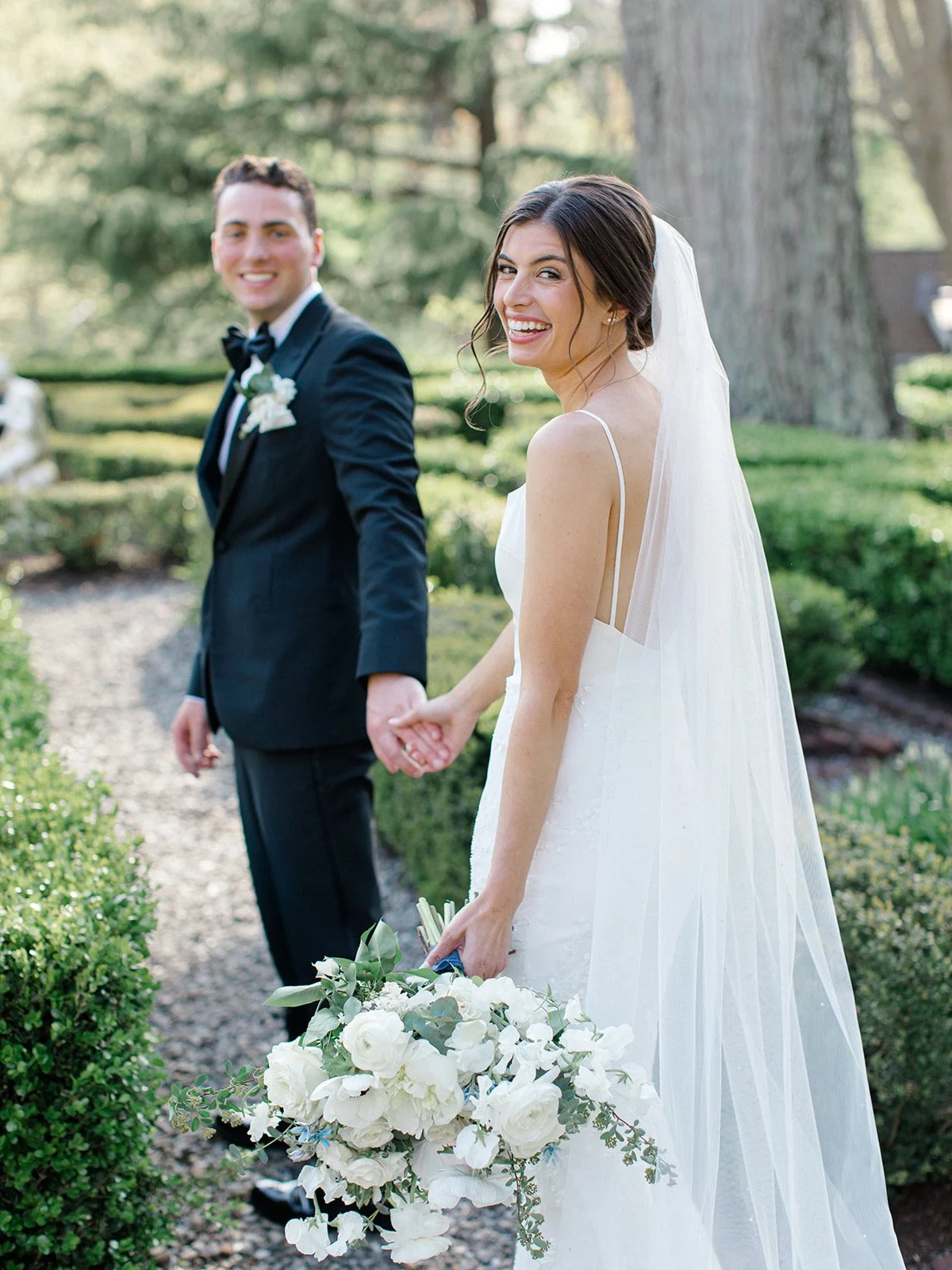 A bride in a white wedding dress smiling and holding a bouquet of white flowers, standing outdoors with a groom in a black tuxedo holding her hand.