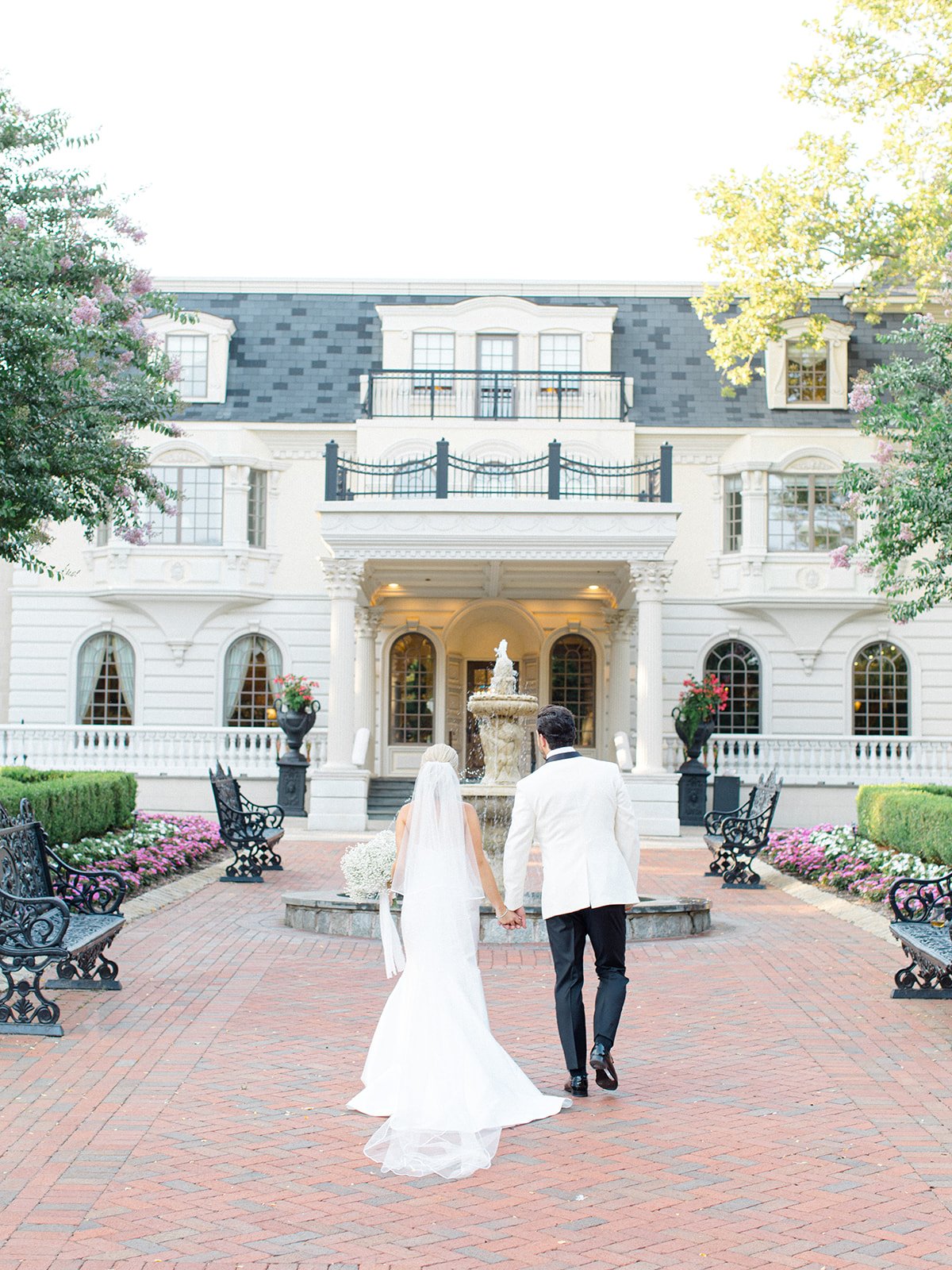 A bride and groom walking hand-in-hand towards a grand white mansion with a fountain in front, surrounded by landscaped gardens and benches.