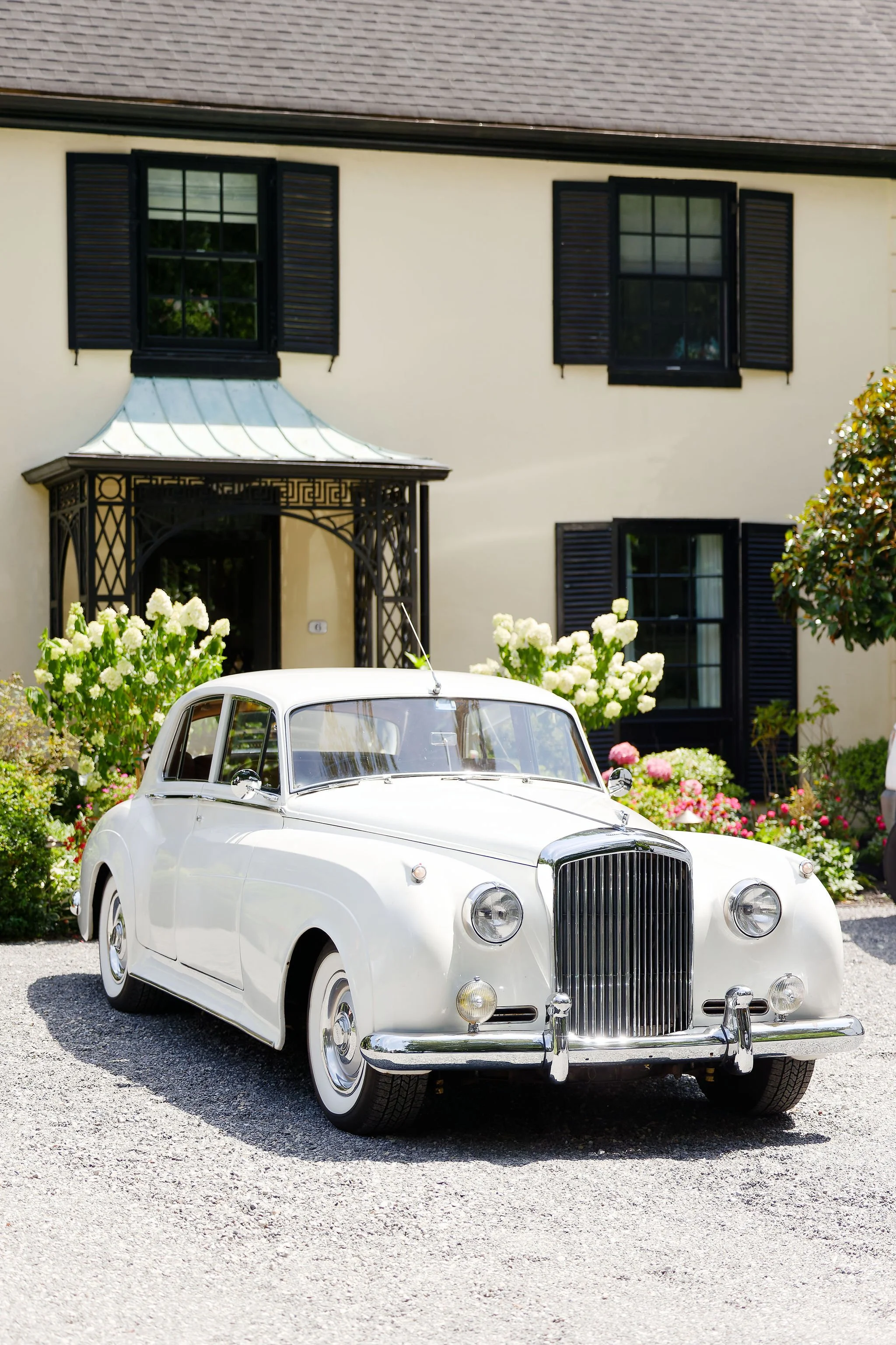 A vintage white Bentley car parked in front of a yellow house with black shutters and a small porch, surrounded by white and pink flowers.