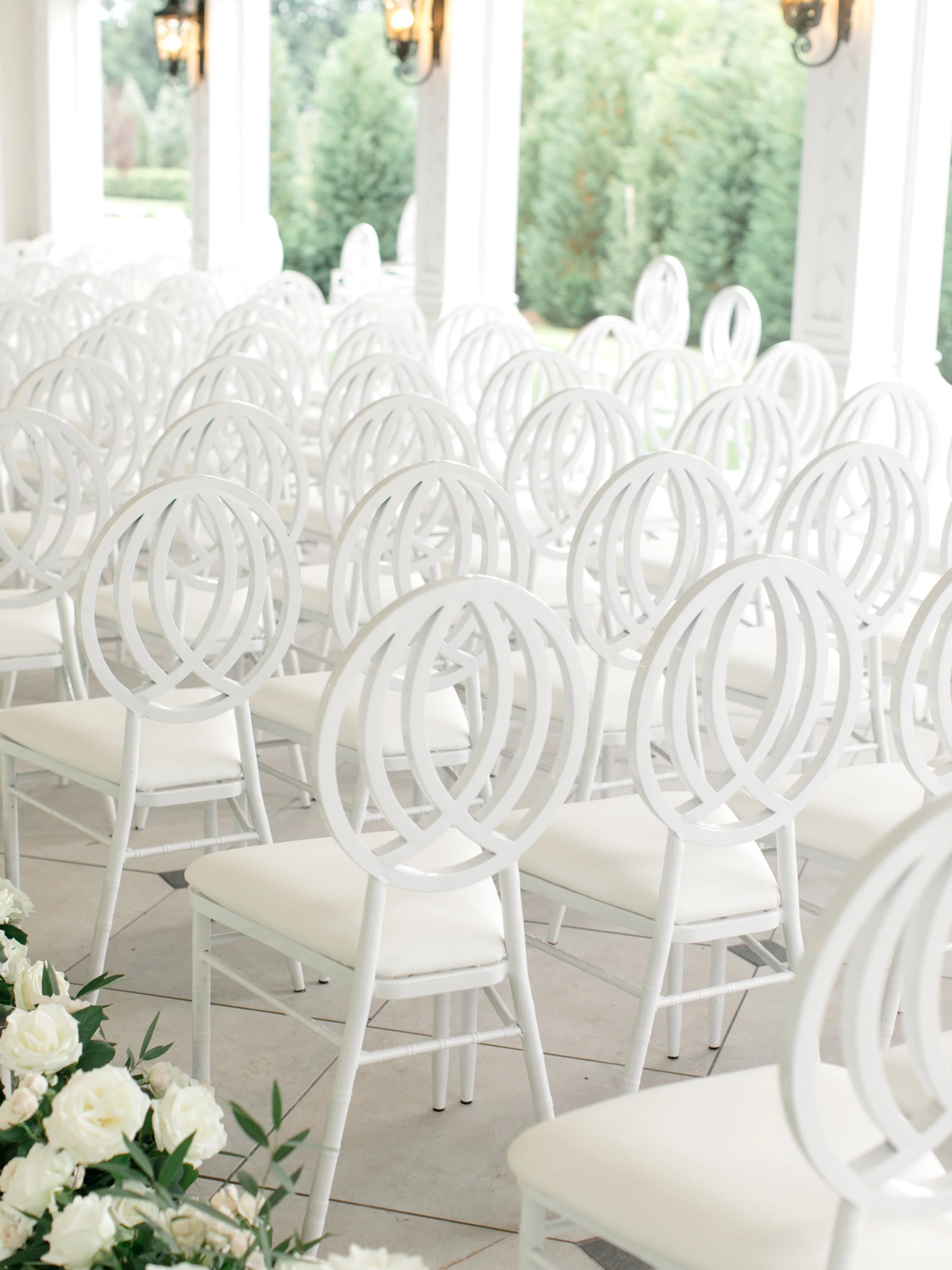 Empty white chairs arranged in rows inside a bright room with large windows overlooking green trees, likely set up for a wedding or event.