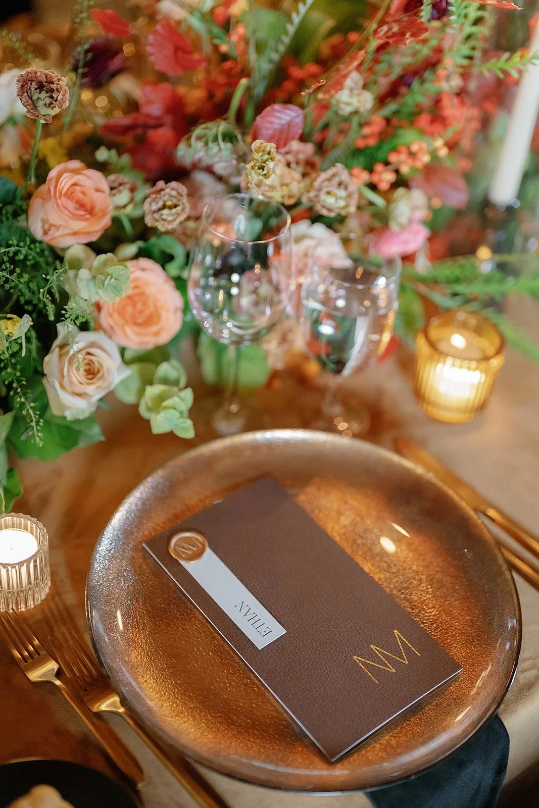 A formal dining table setup with a metallic gold plate, gold utensils, a leather-bound menu labeled 'EULIAN,' a glass of water, a wine glass, lit candles in gold holders, and a centerpiece of mixed pink and red flowers and greenery.