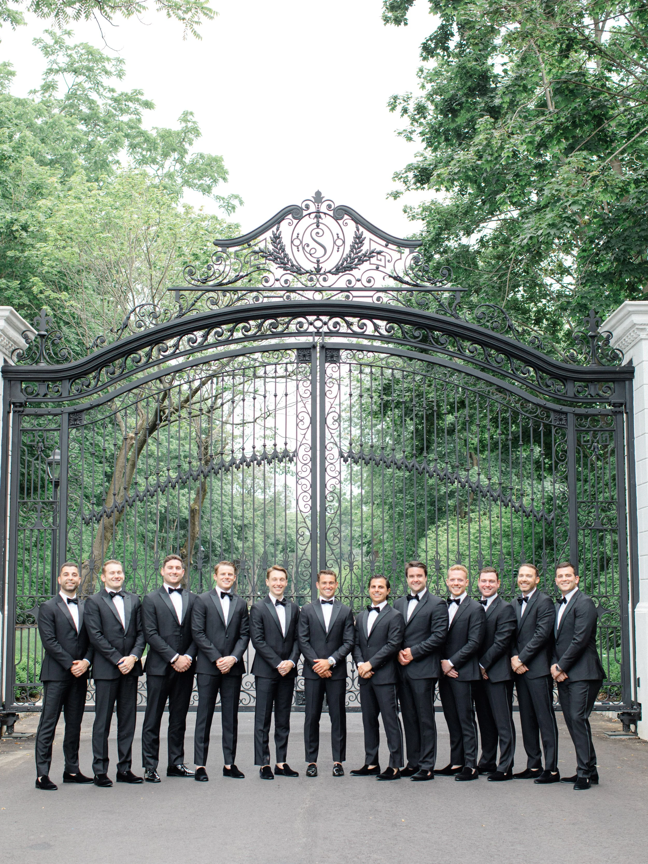 A group of fourteen men dressed in black tuxedos with bow ties standing in front of a large ornate wrought iron gate with trees in the background.